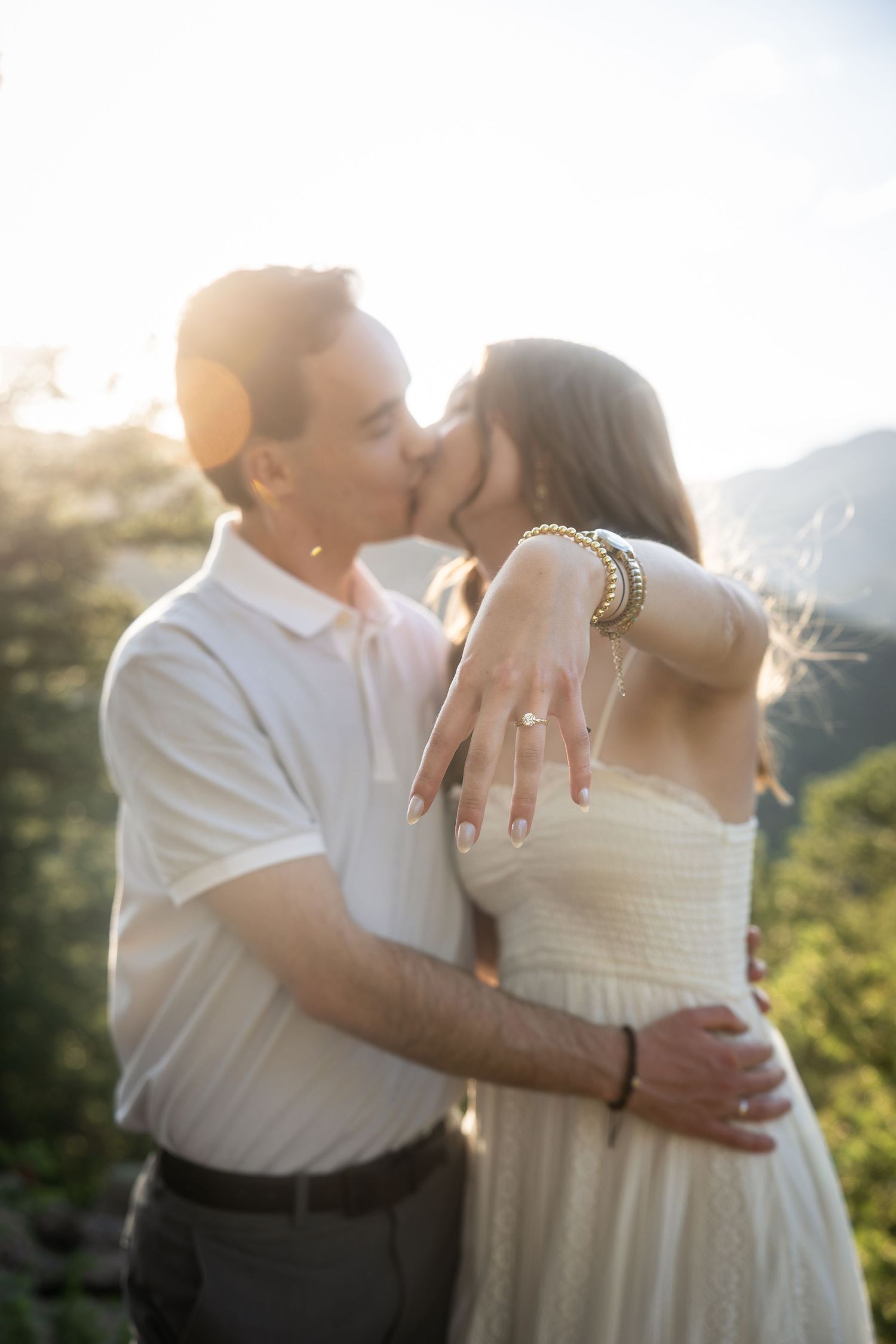 Couple kissing at sunset outdoors, woman displaying engagement ring on outstretched hand with mountains in background.