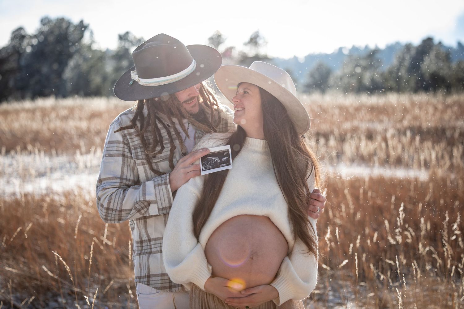 Couple embraces in a golden wheat field on a sunny afternoon wearing neutral toned clothing.