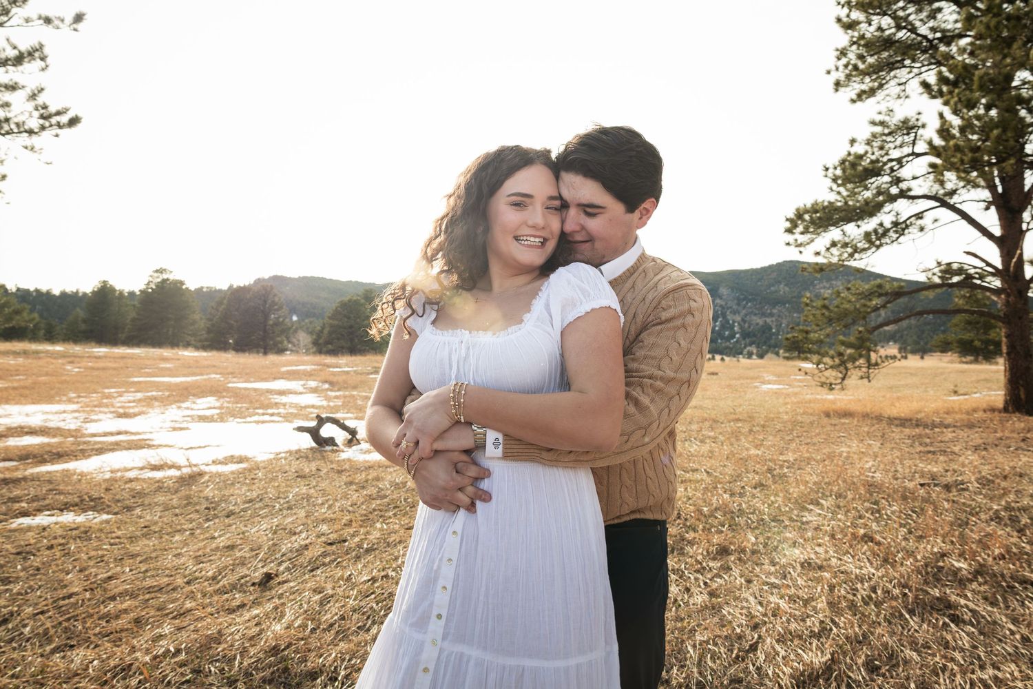 Couple embracing in golden field with mountains and trees at sunset, man in tan sweater hugging woman in white dress.