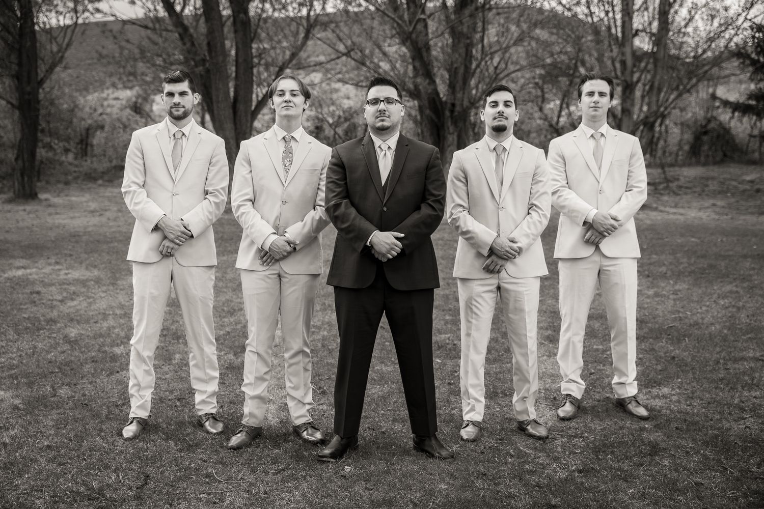 Black and white photo of a groom in dark suit flanked by four groomsmen in light suits standing outdoors.