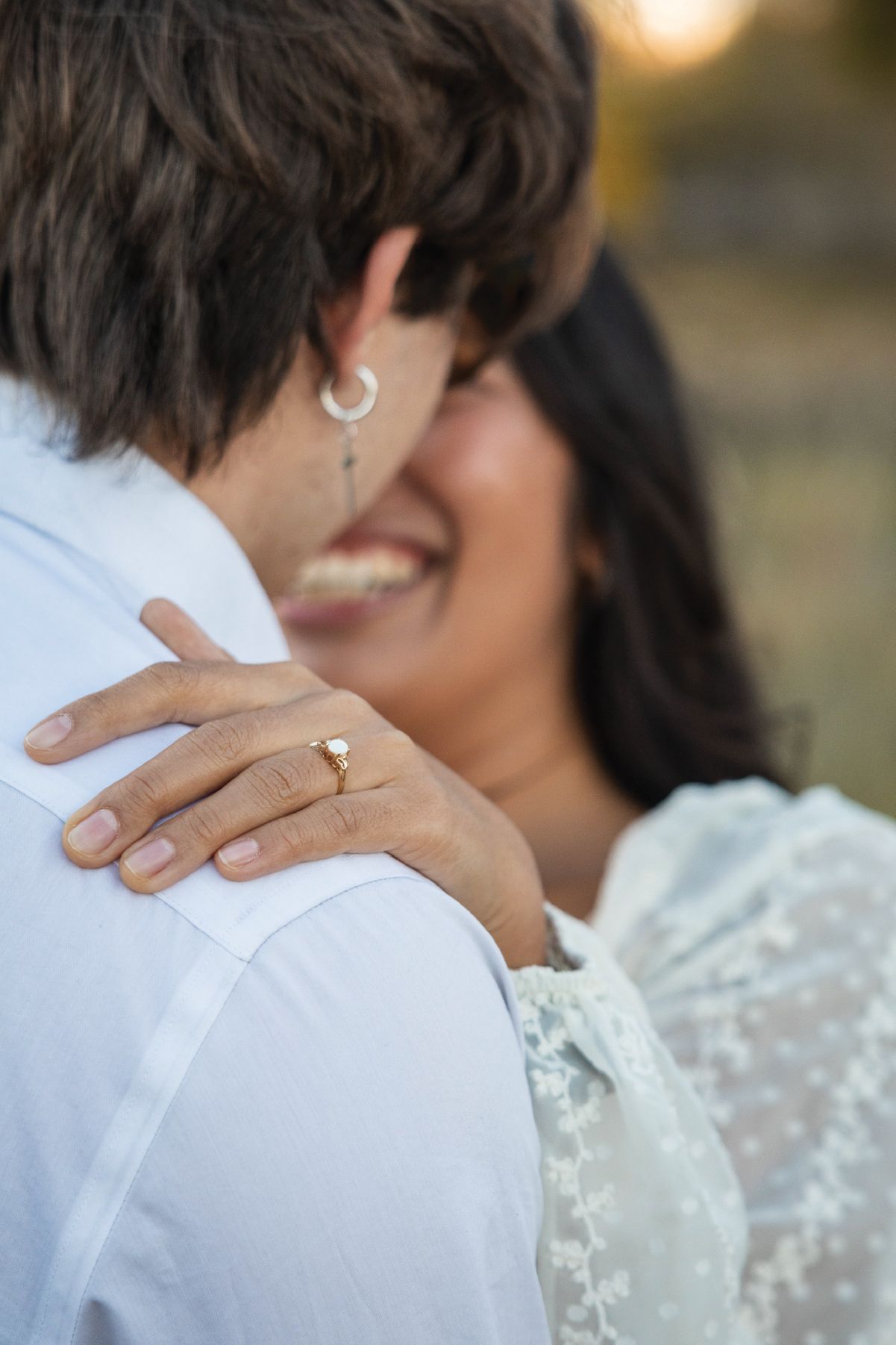 A joyful woman in white lace dress embraces her partner, showcasing a gold engagement ring on her hand.
