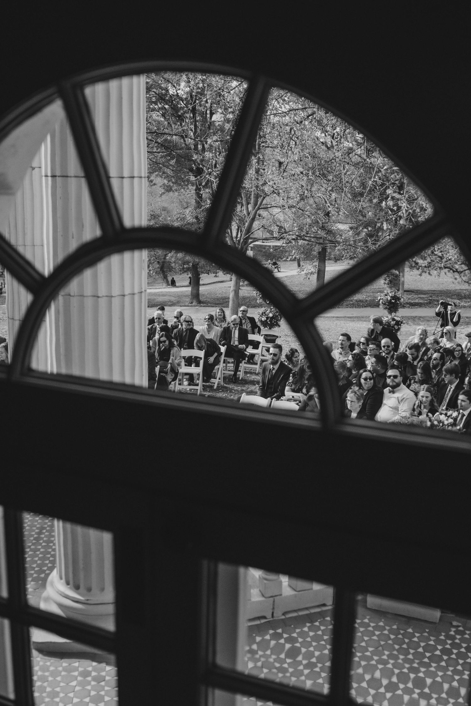 Black and white view through an arched window showing outdoor wedding ceremony seating.