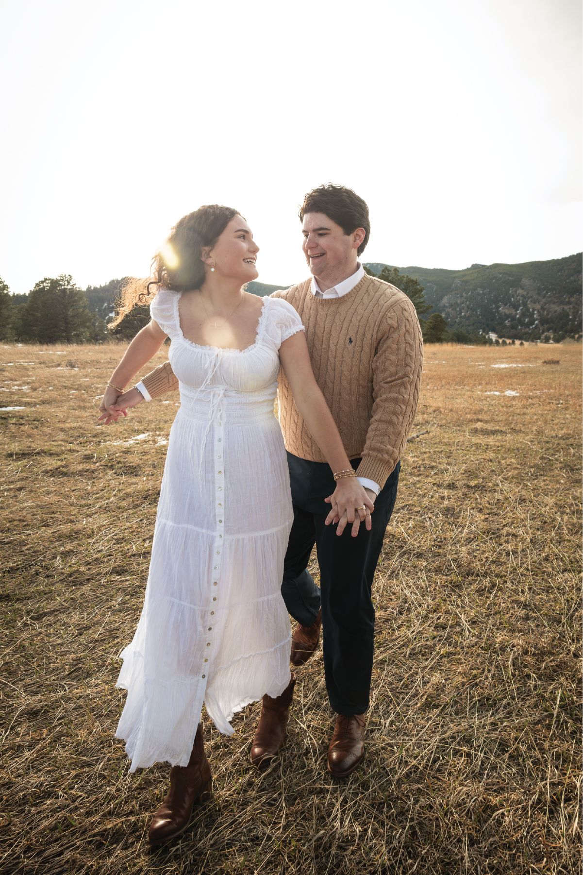 A couple walks hand-in-hand through a golden field at sunset, smiling at each other in white dress and tan sweater.