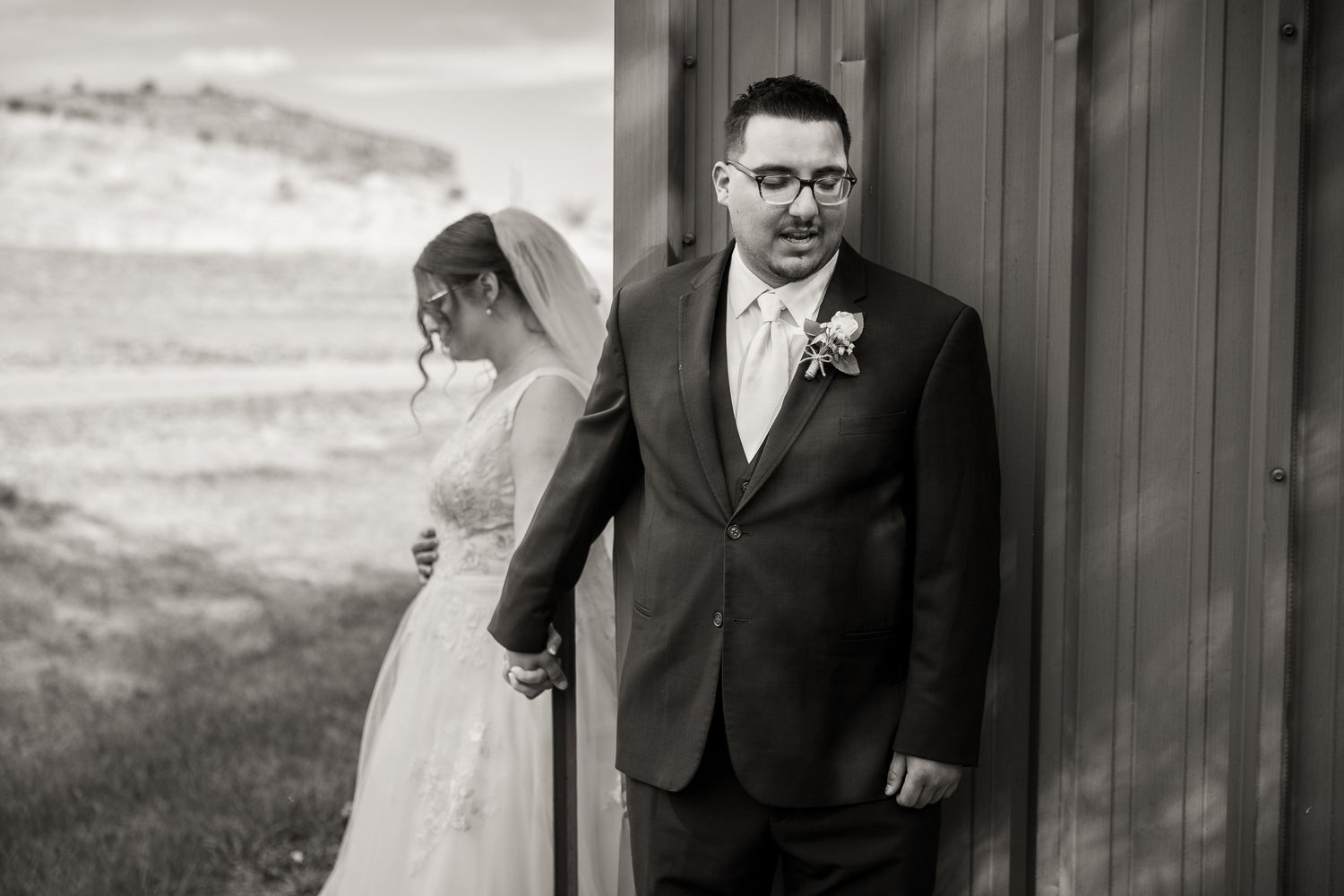 Black and white photo of a bride and groom sharing a first look moment beside a rustic barn wall.