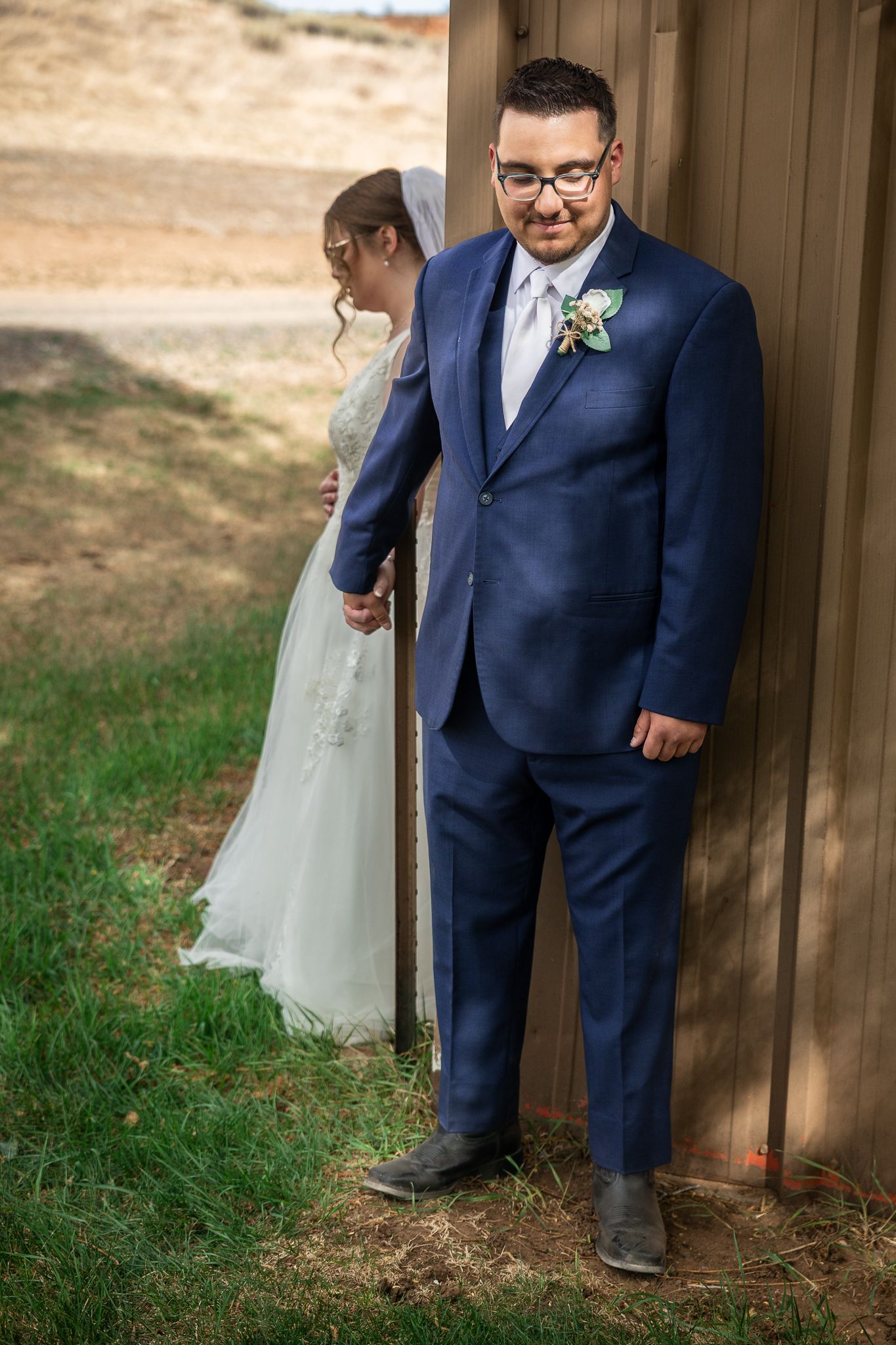 Groom in blue suit with boutonniere stands by wooden wall as bride in white gown peeks around corner.