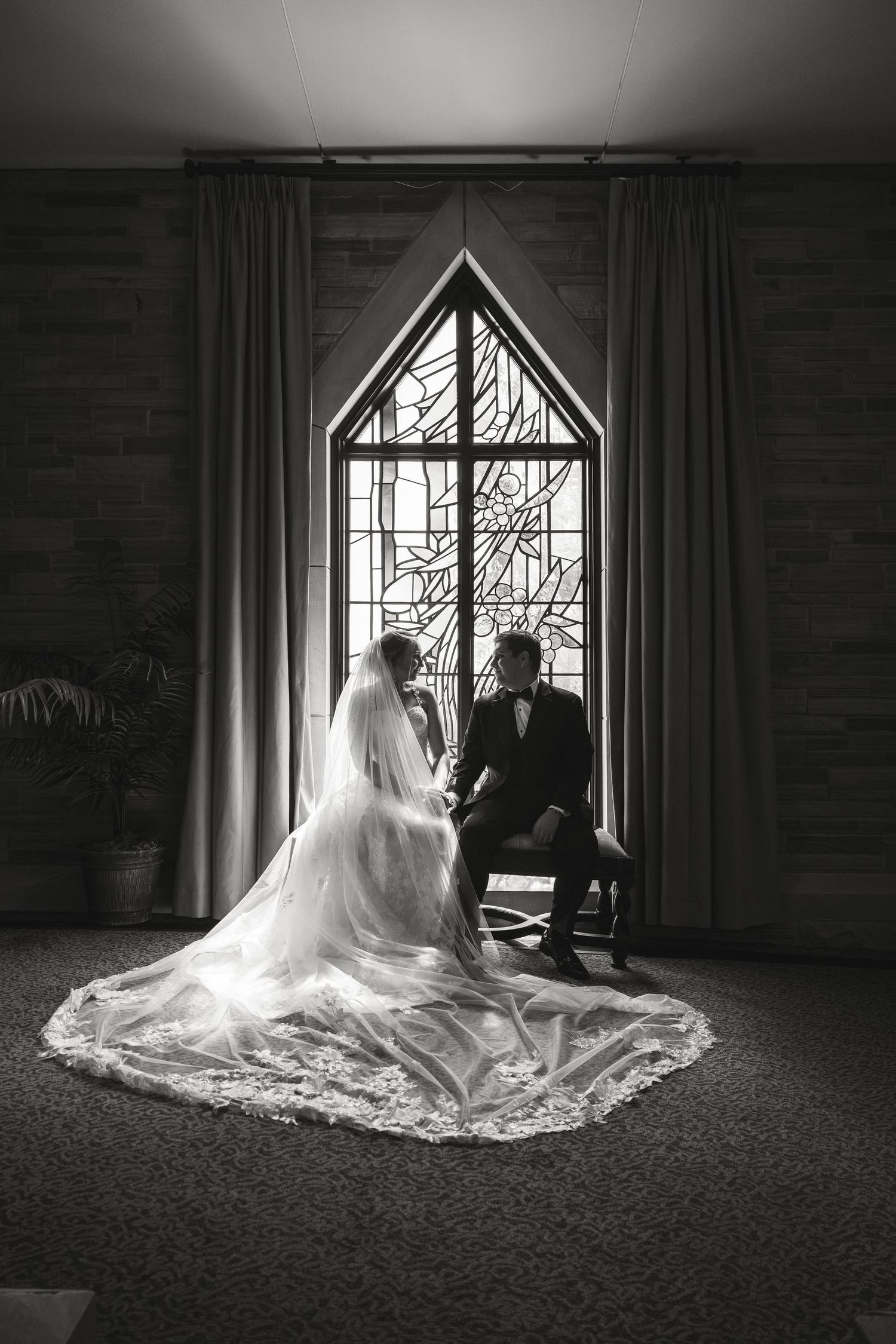 Black and white photo of bride and groom sitting by a stained glass chapel window, bridal veil spread elegantly on floor.