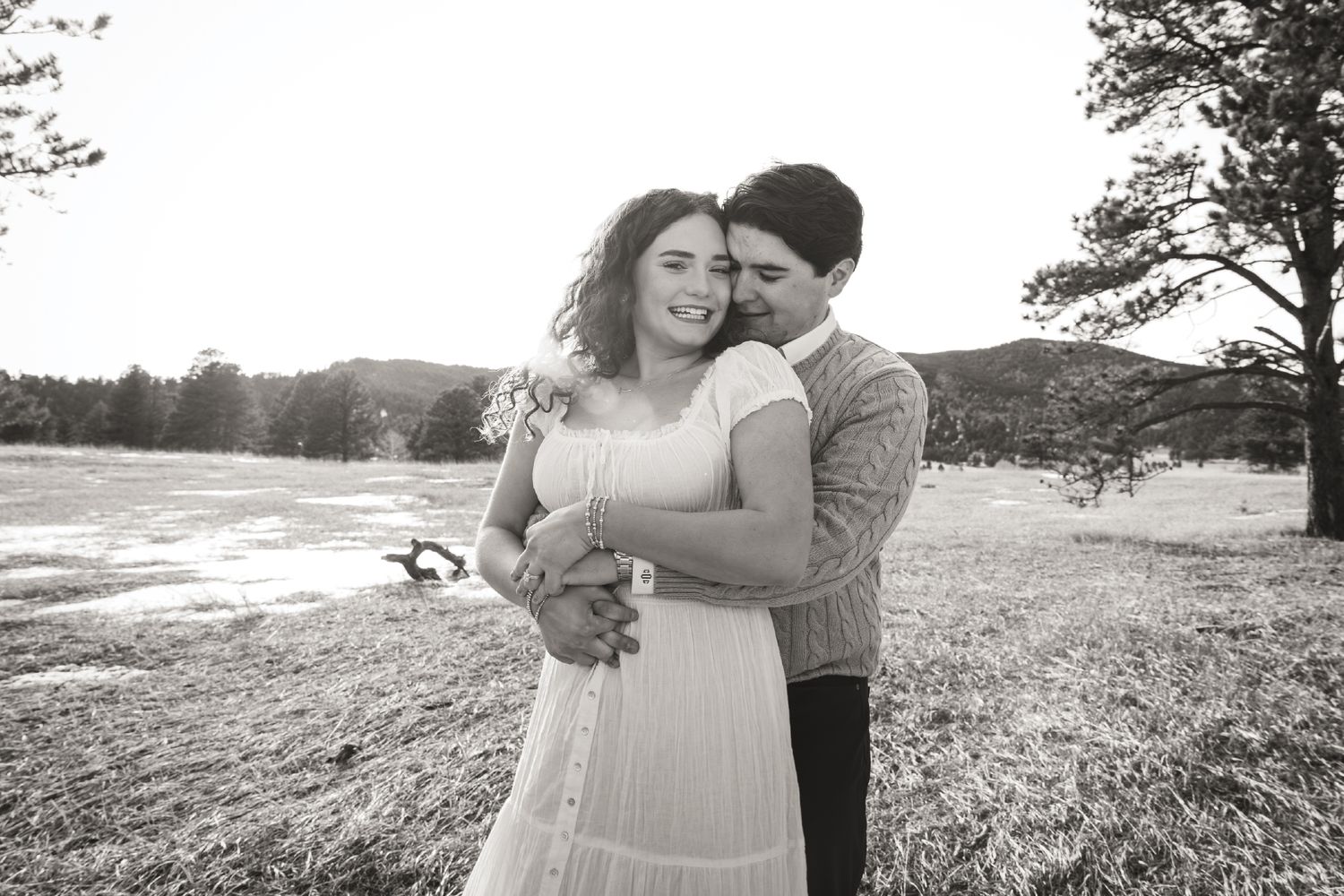Black and white photo of a couple embracing in a scenic outdoor field with trees and mountains in the background.