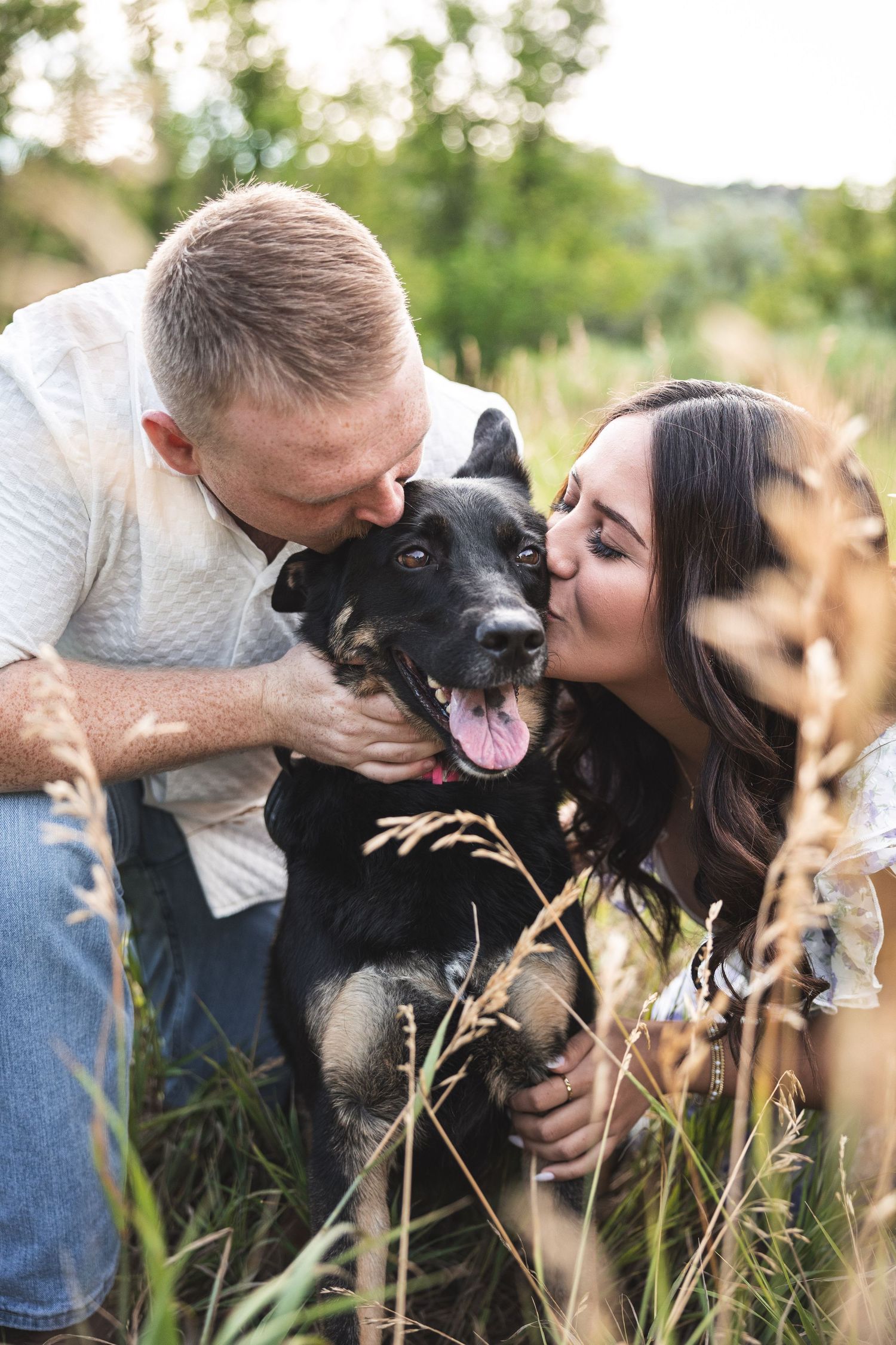 Couple kissing their happy black dog in a lush green field, capturing a joyful outdoor pet photography moment.