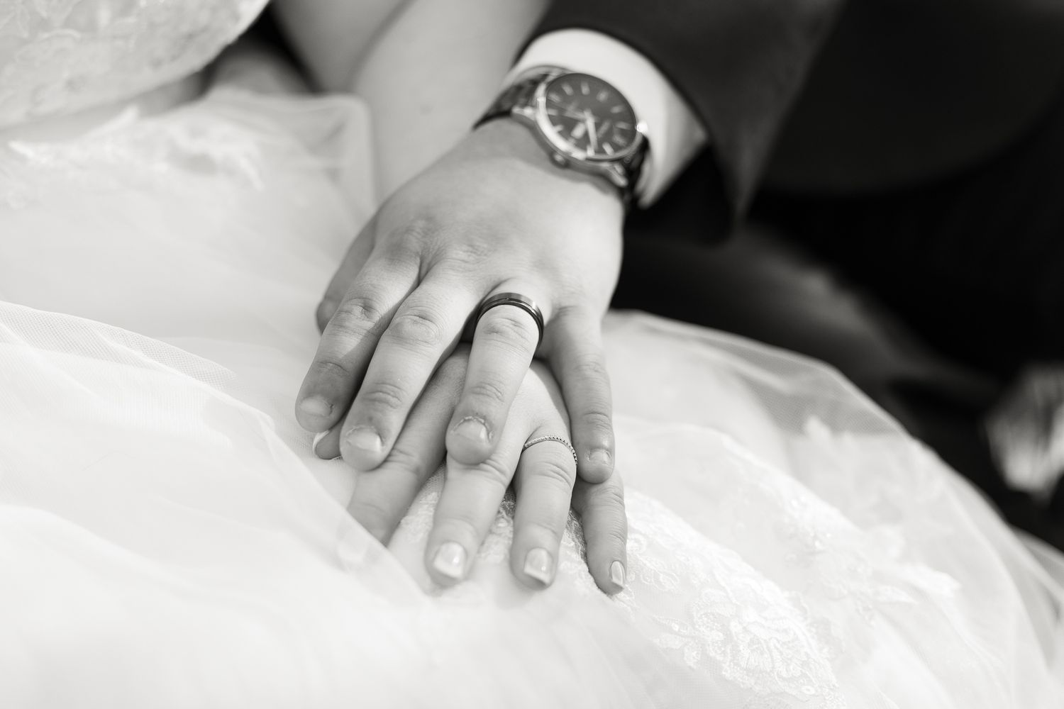 Black and white wedding photo of bride and groom hands together showing rings on white dress.