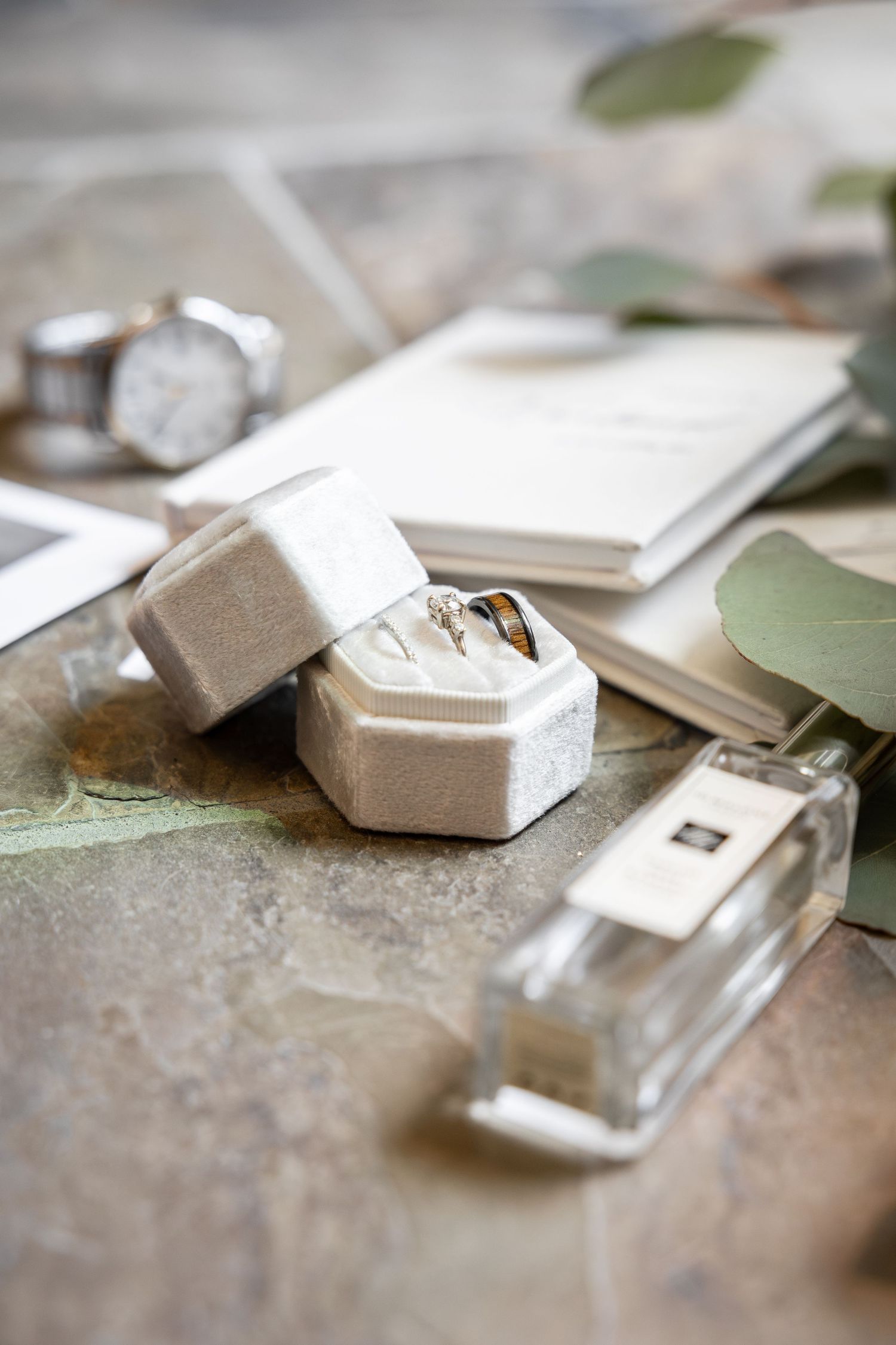 Wedding rings in velvet box with watch, perfume, and eucalyptus on stone surface flat lay detail shot.