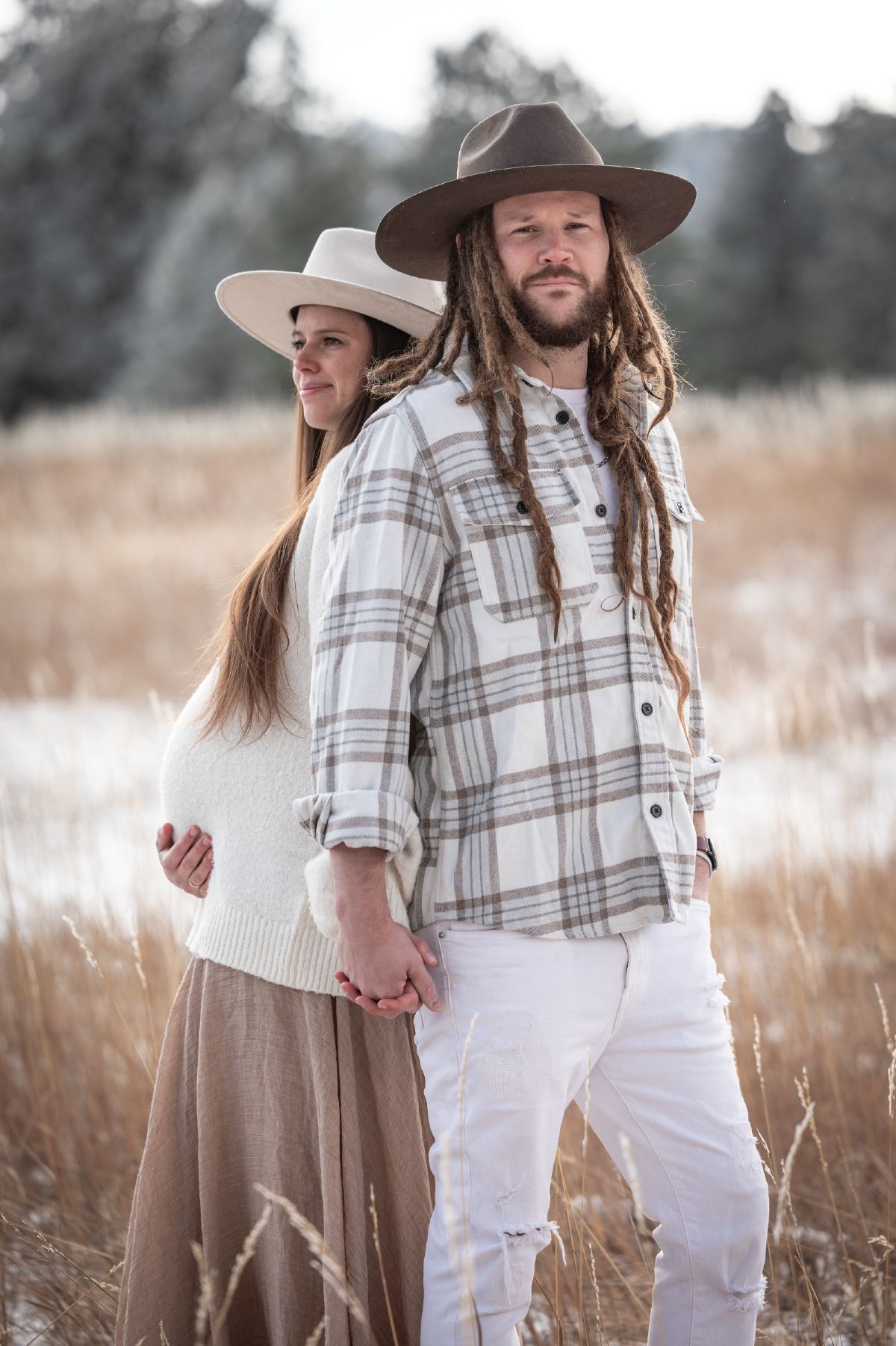 Couple in wide-brim hats holding hands in golden field, man with dreadlocks in plaid shirt, woman in cream sweater.