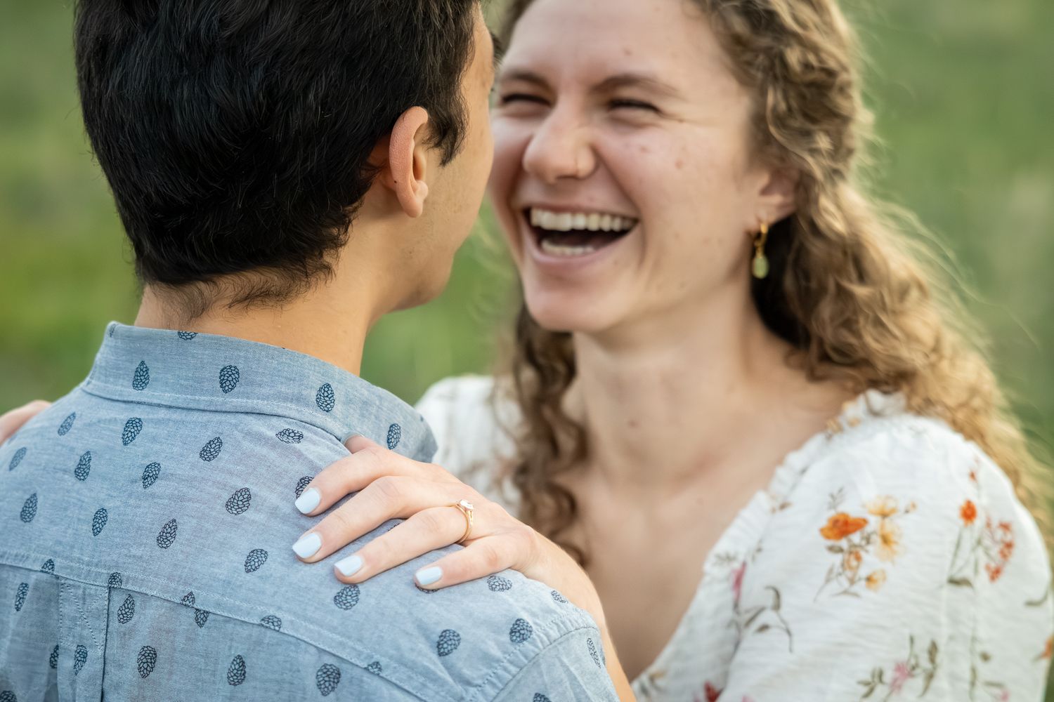 A laughing woman in a floral dress rests her hand on a man's shoulder during a romantic outdoor engagement photo.