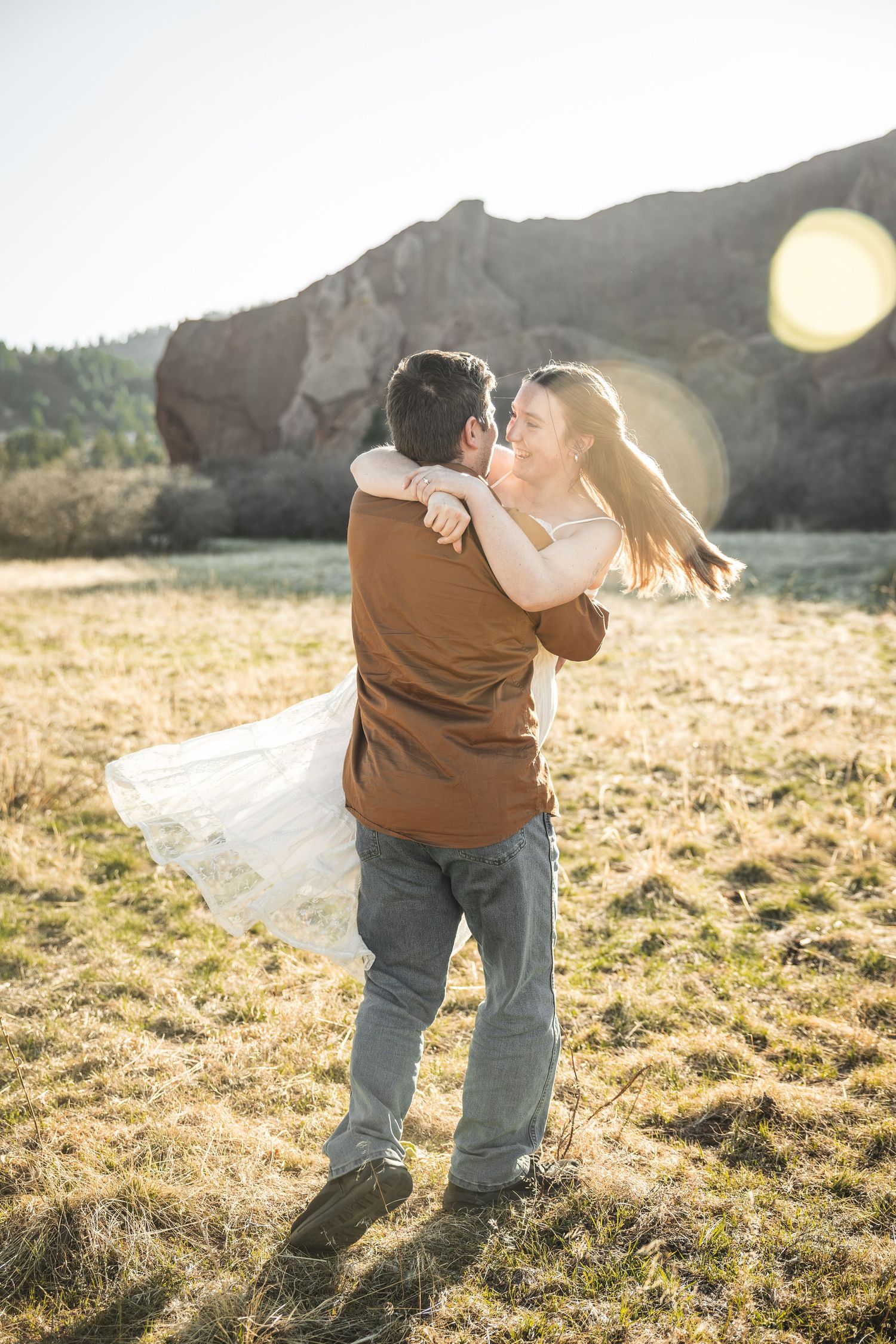 A groom lifts his bride in a sunlit field with rocky mountains in the background during a golden hour outdoor engagement shoot.
