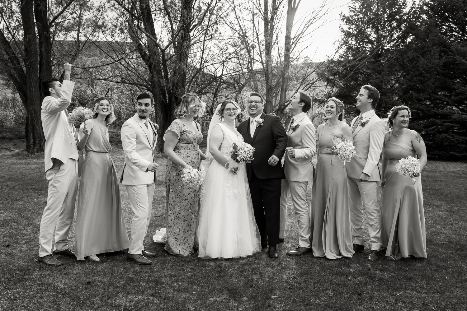 Black and white wedding party photo with bride, groom, and attendants posing outdoors on grass near trees.