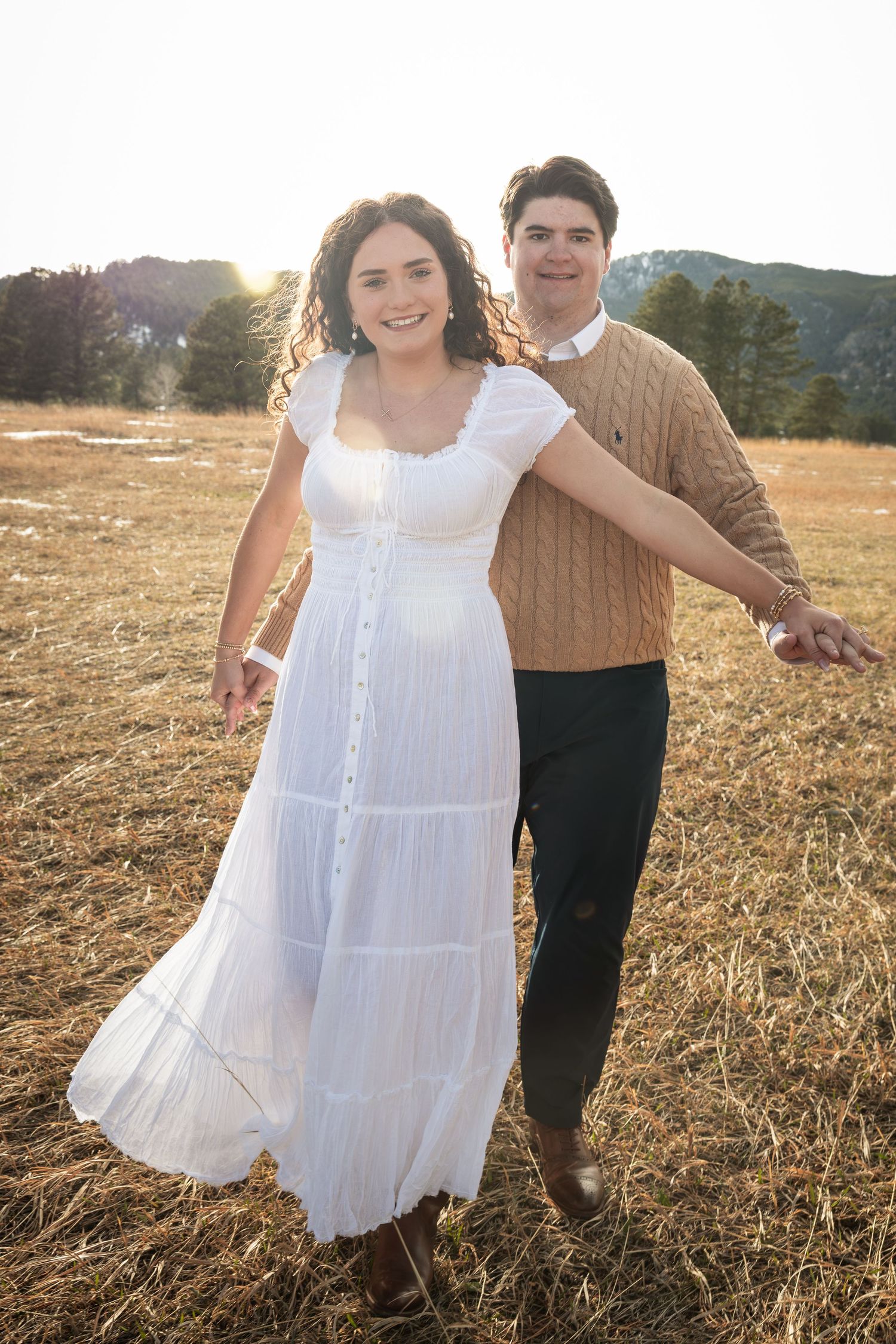 Couple walking hand-in-hand in a sunlit field, woman in white dress, man in tan sweater, mountains in background.