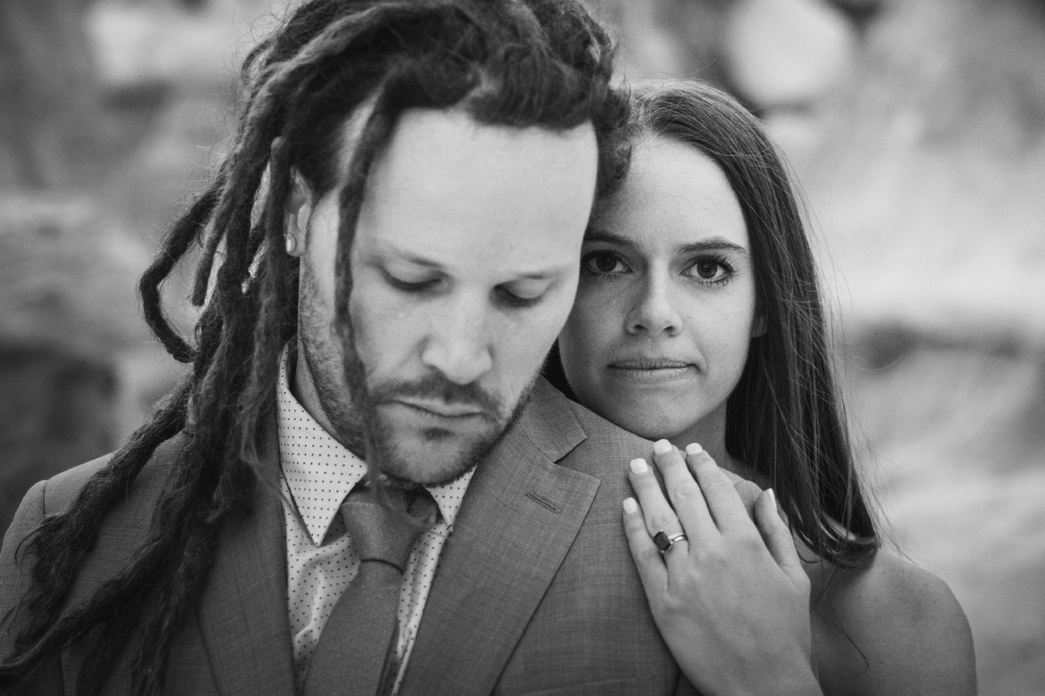 Black and white portrait of a couple, man with dreadlocks in suit, woman showing engagement ring, romantic pose.
