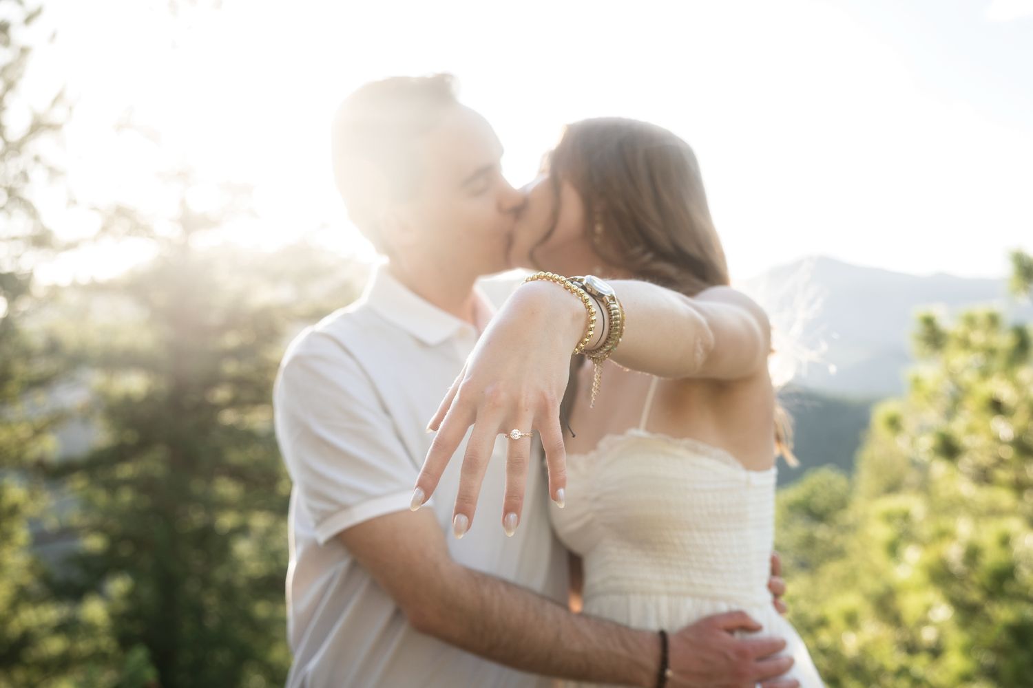 Couple kissing at sunset in mountains, woman showing engagement ring, wearing white outfits with golden bracelet.