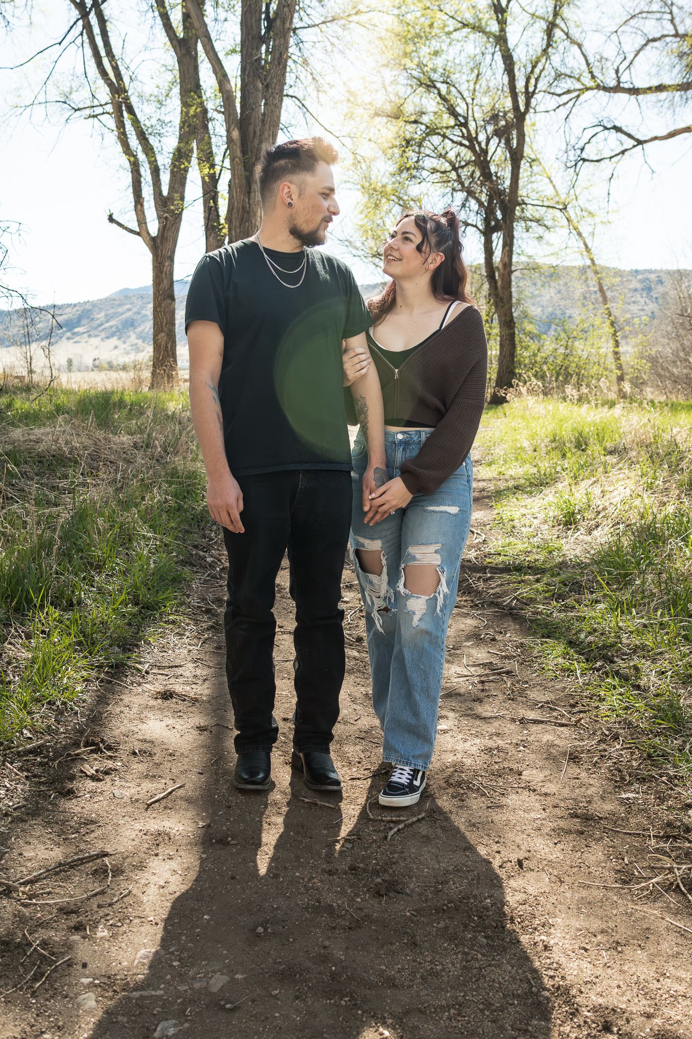 A young couple holds hands on a dirt path surrounded by trees and greenery on a sunny day.