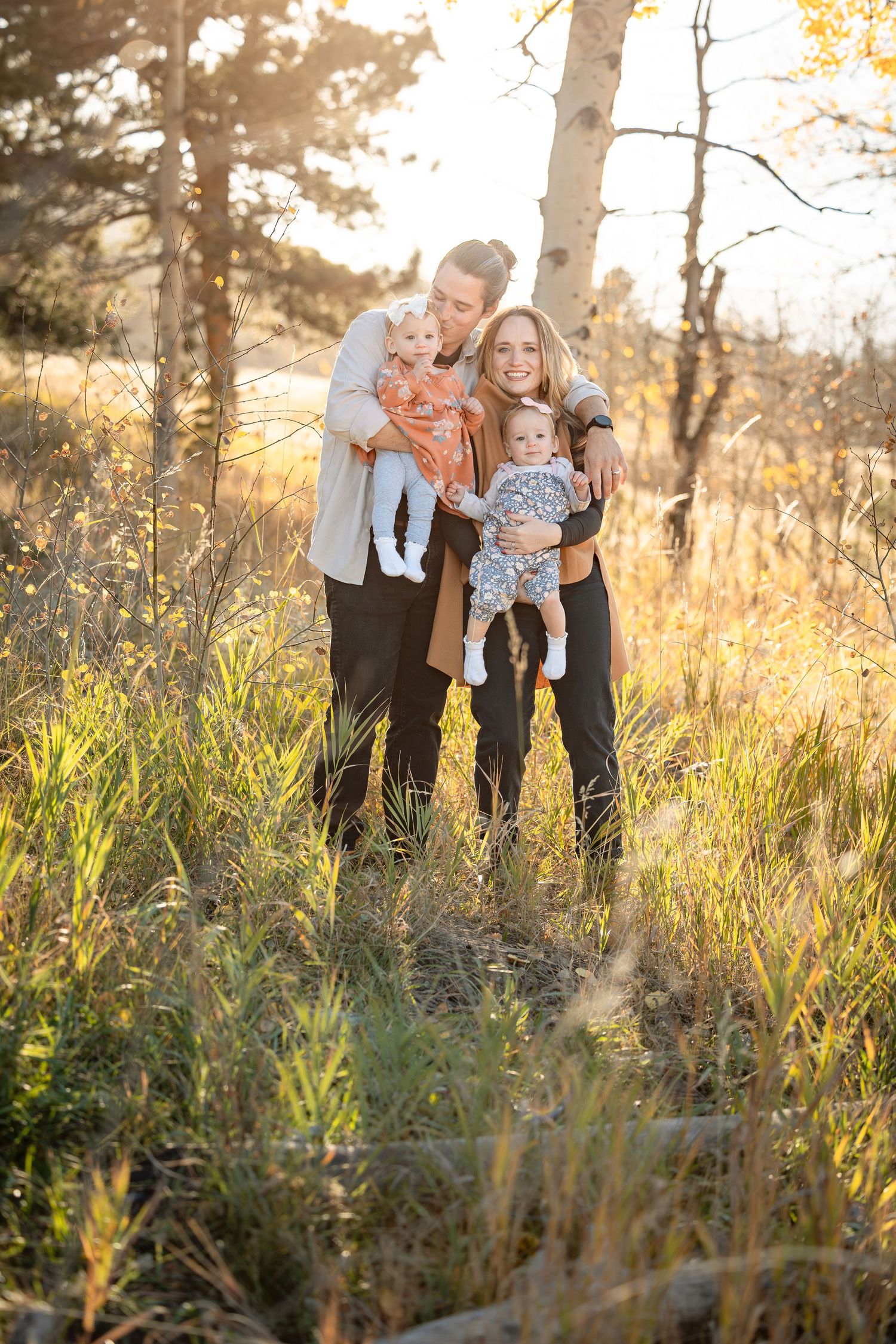 Two women holding babies smile outdoors in golden autumn light among tall grass and trees.