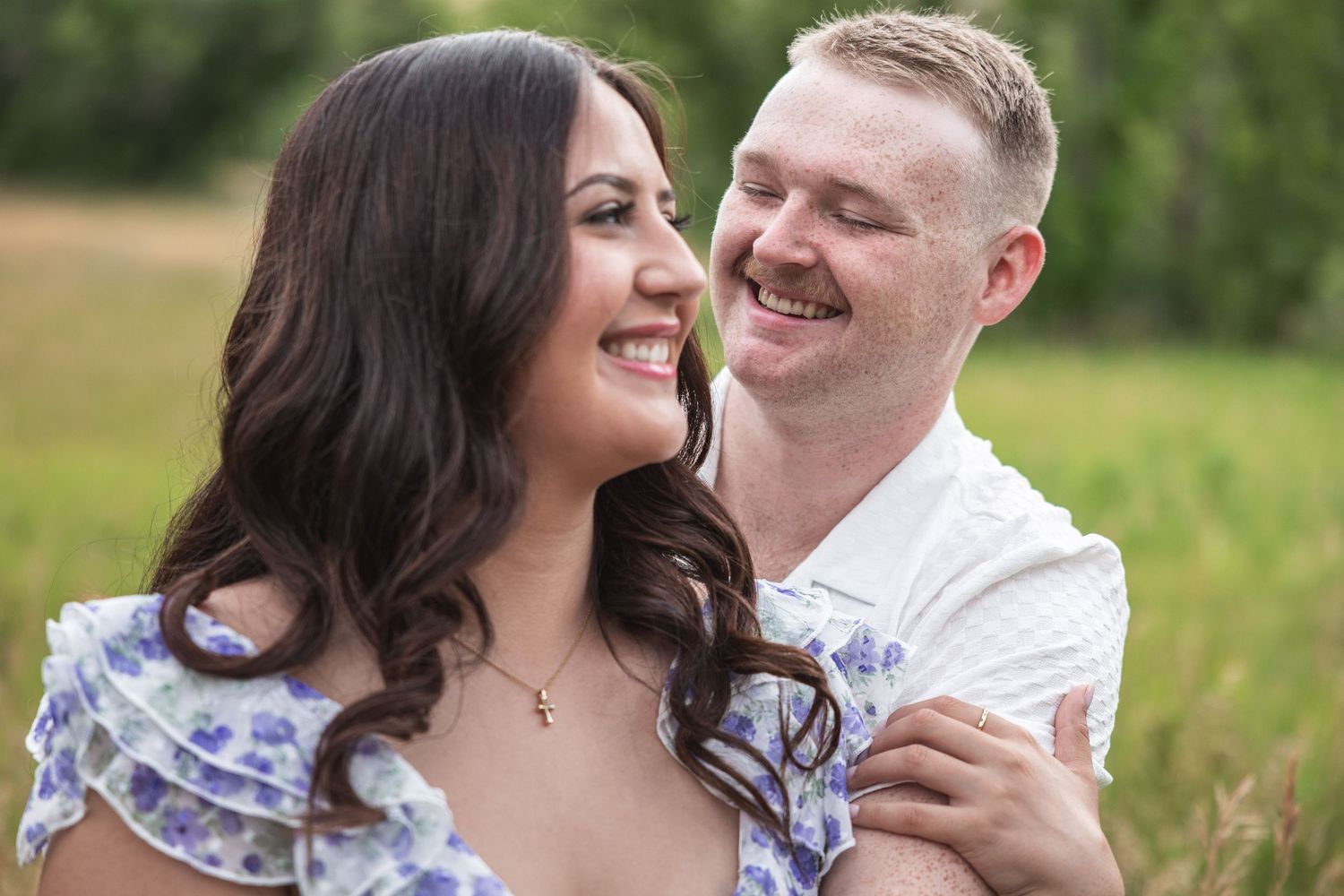 A smiling couple shares a tender moment outdoors, woman in floral dress, man in white shirt, green field background.