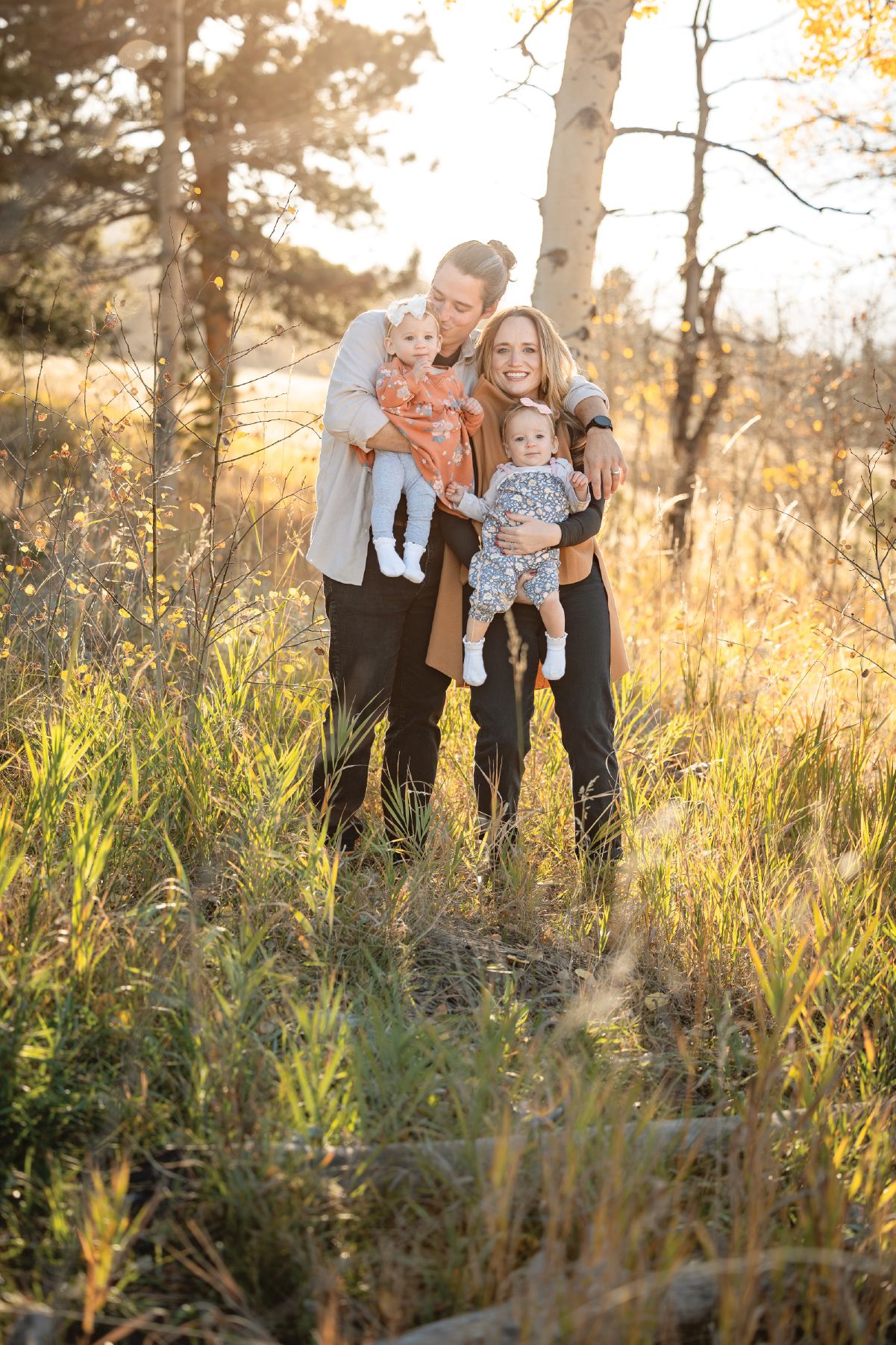 Happy family of four poses in golden sunlit meadow, parents holding two babies among tall grass and autumn trees.
