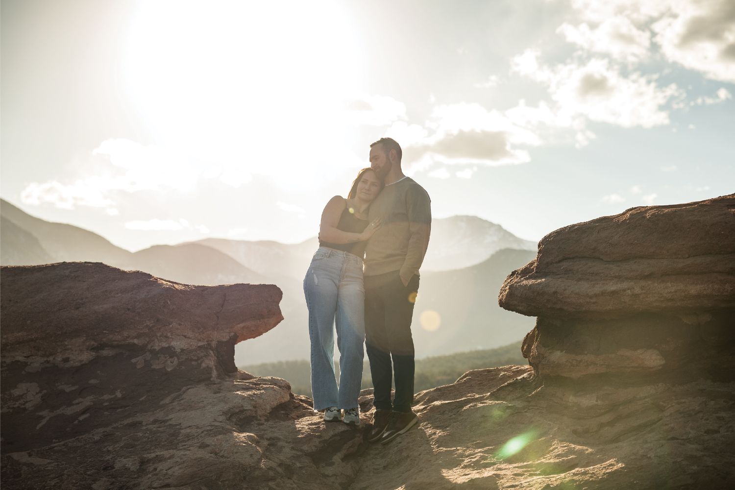 Couple embracing on rocky mountain summit with stunning valley views and golden sunlight in the background.