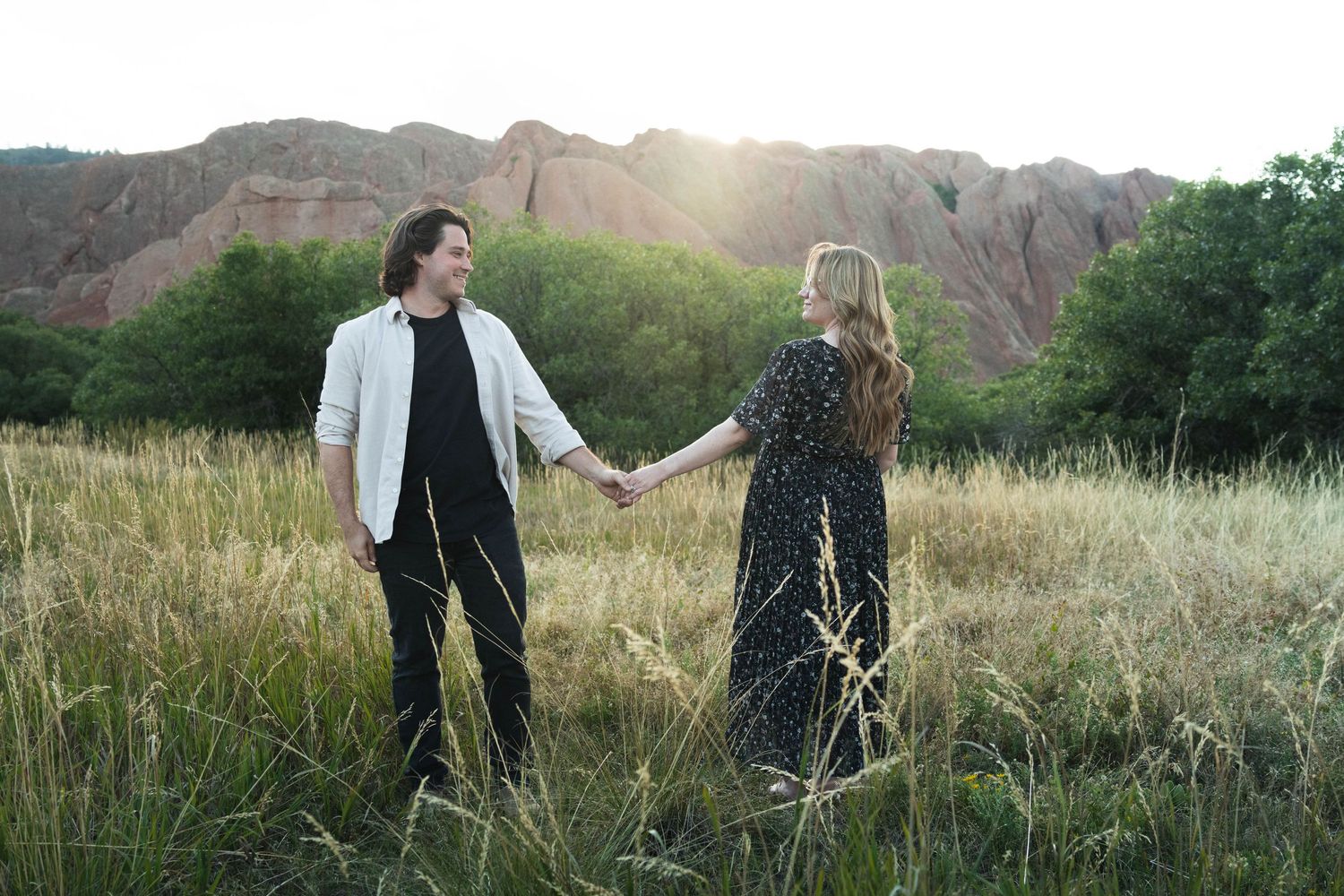 Couple holding hands in golden field with red rock formations and glowing sunset backdrop.