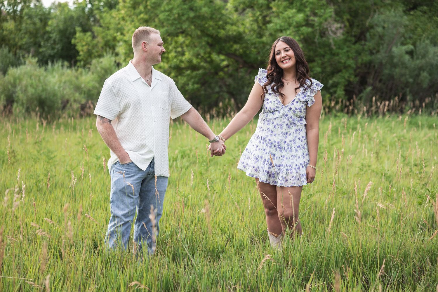 Couple holding hands and smiling while walking through a lush green meadow, woman in floral dress and man in white shirt.