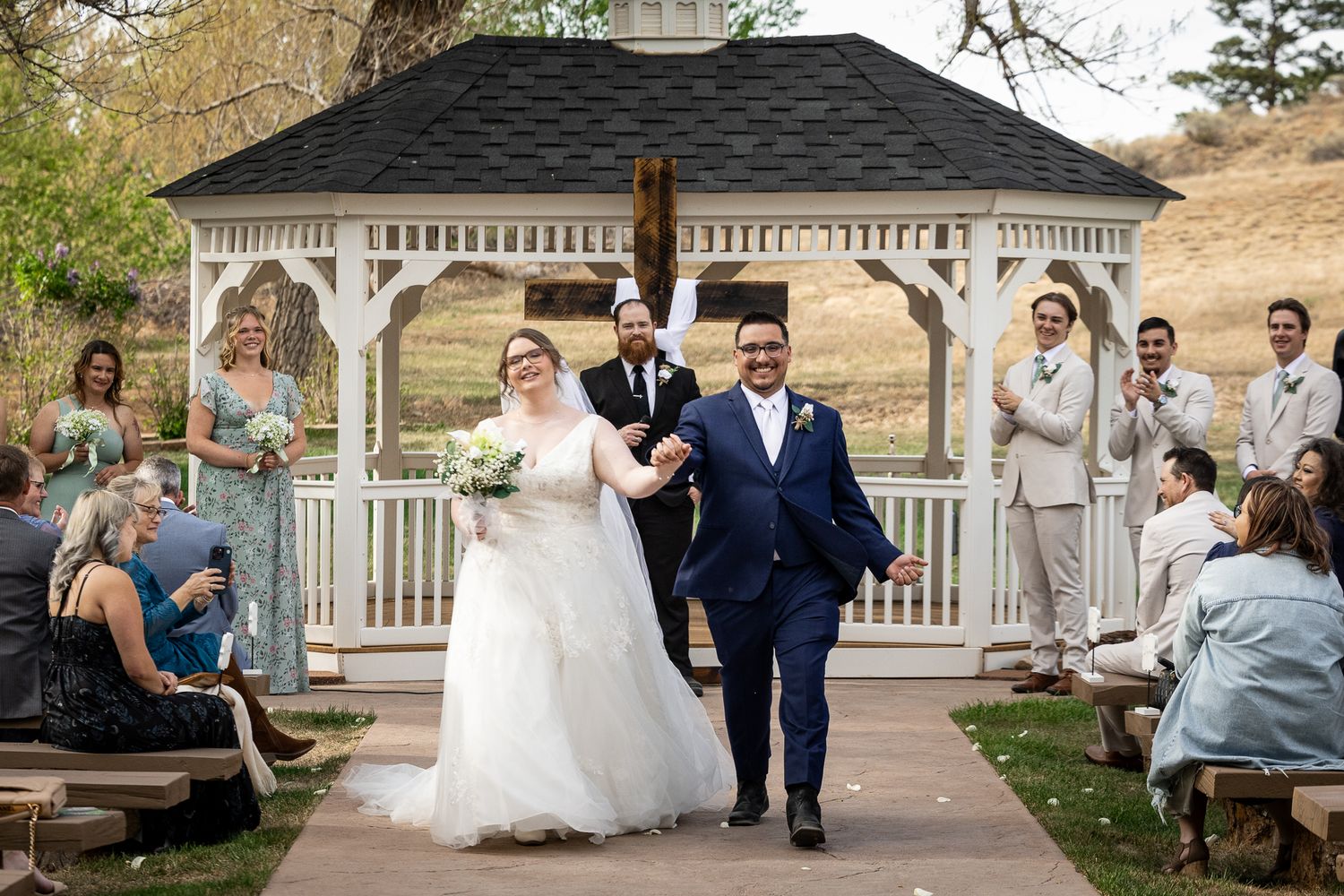 Newlywed couple walks down aisle after outdoor wedding ceremony near white gazebo, surrounded by cheering guests.