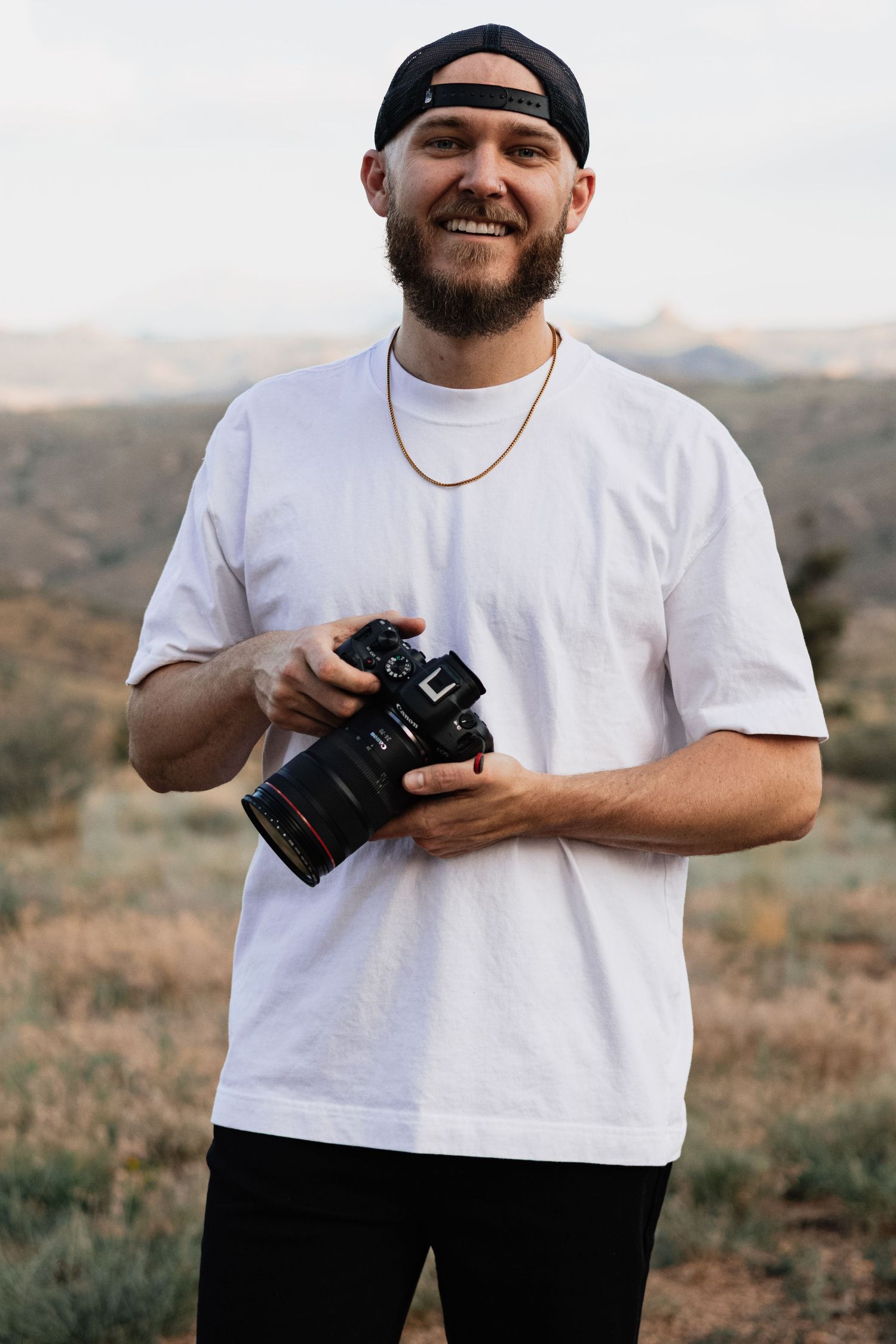 A photographer in a white t-shirt and black pants smiles while holding a camera in an outdoor landscape setting.