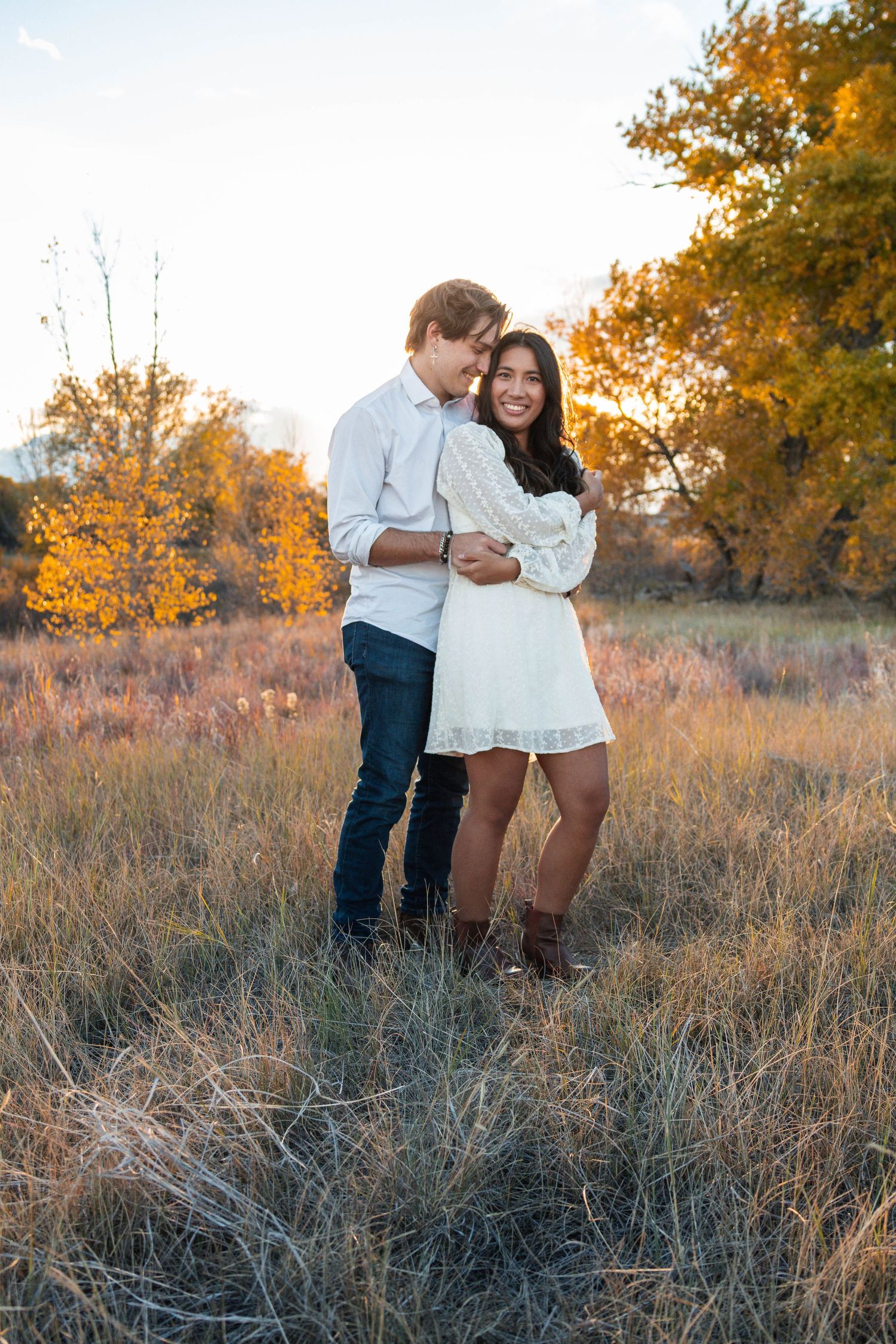 Two people share a romantic moment in a fall meadow with orange trees and dried grass at golden hour.