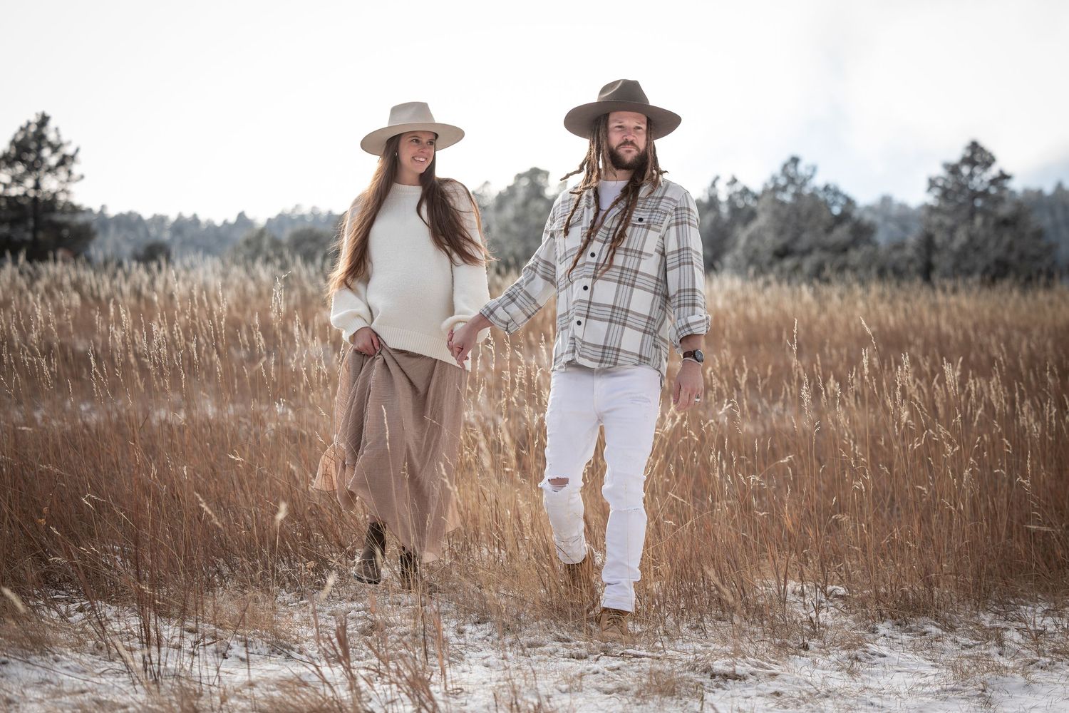 Romantic scene of two people walking through a sunlit field wearing earth-toned outfits and wide-brimmed hats.