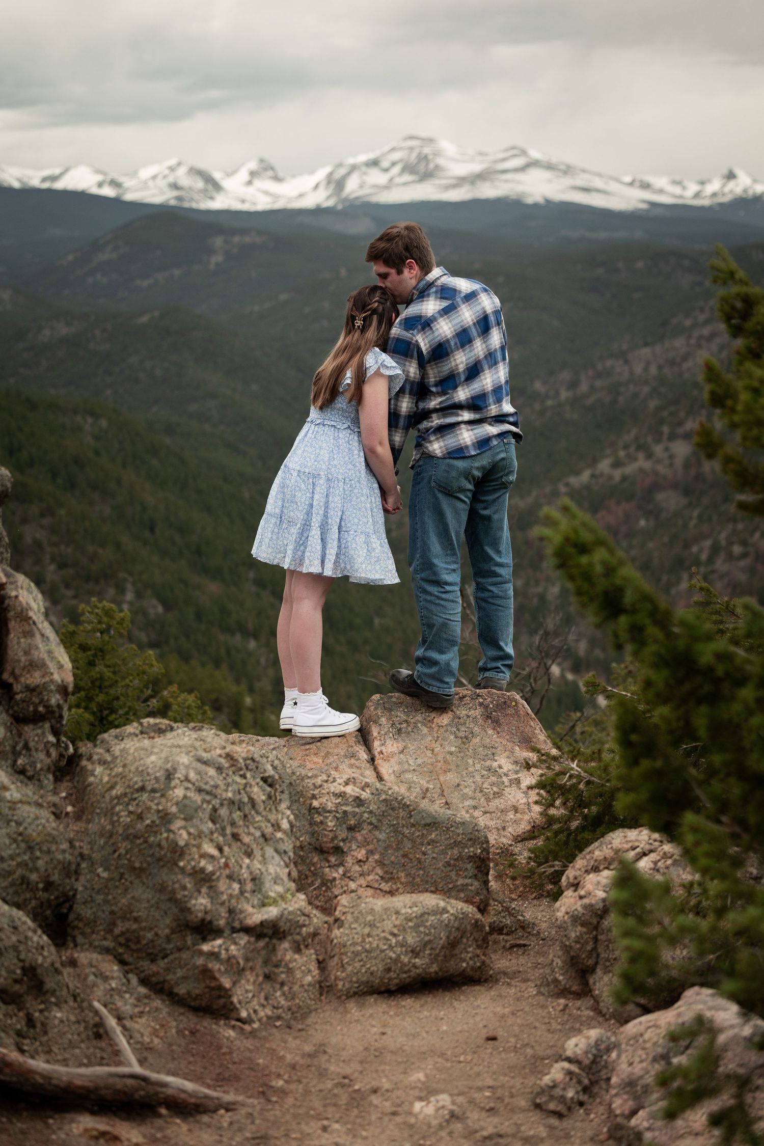 Couple standing on mountain overlook with snow-capped peaks visible in the background.