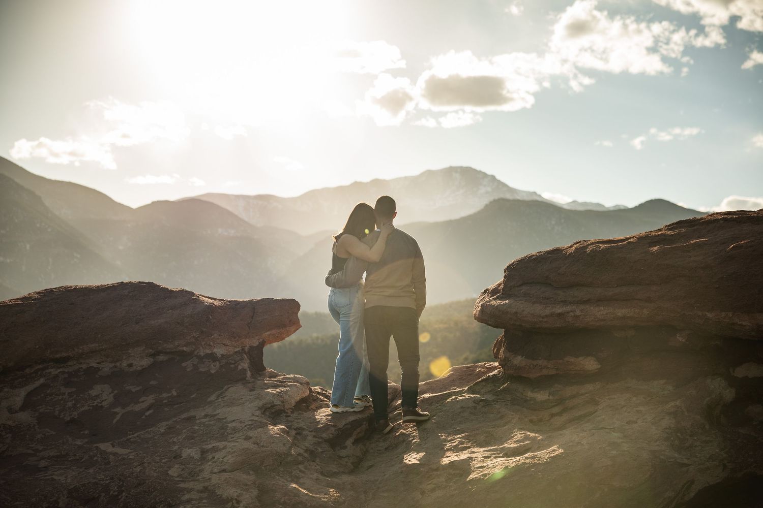 Silhouettes of two people sitting on rocky terrain against a mountainous sunset backdrop.