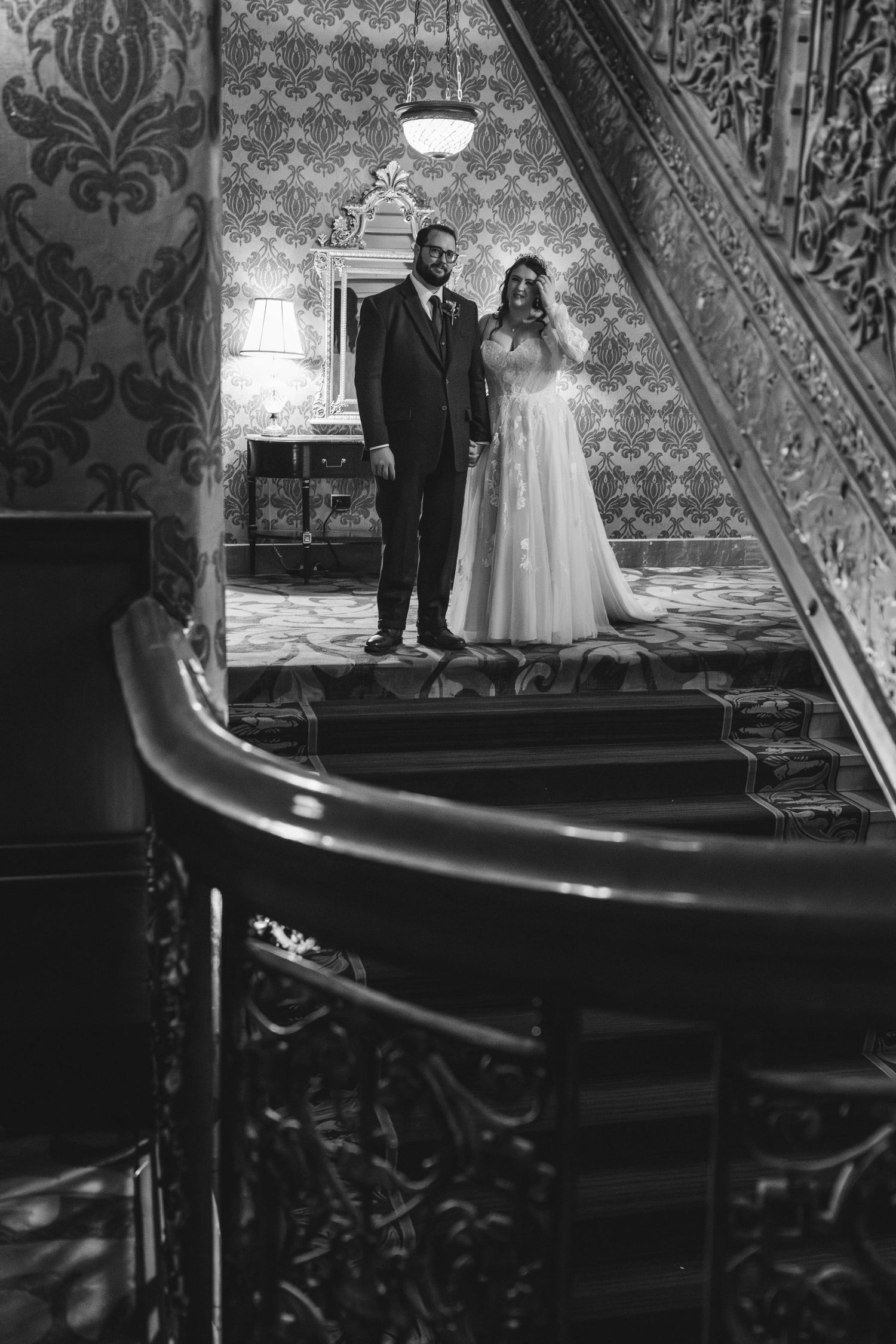 A wedding couple poses on vintage stairs with ornate wallpaper and detailed banister in black and white.