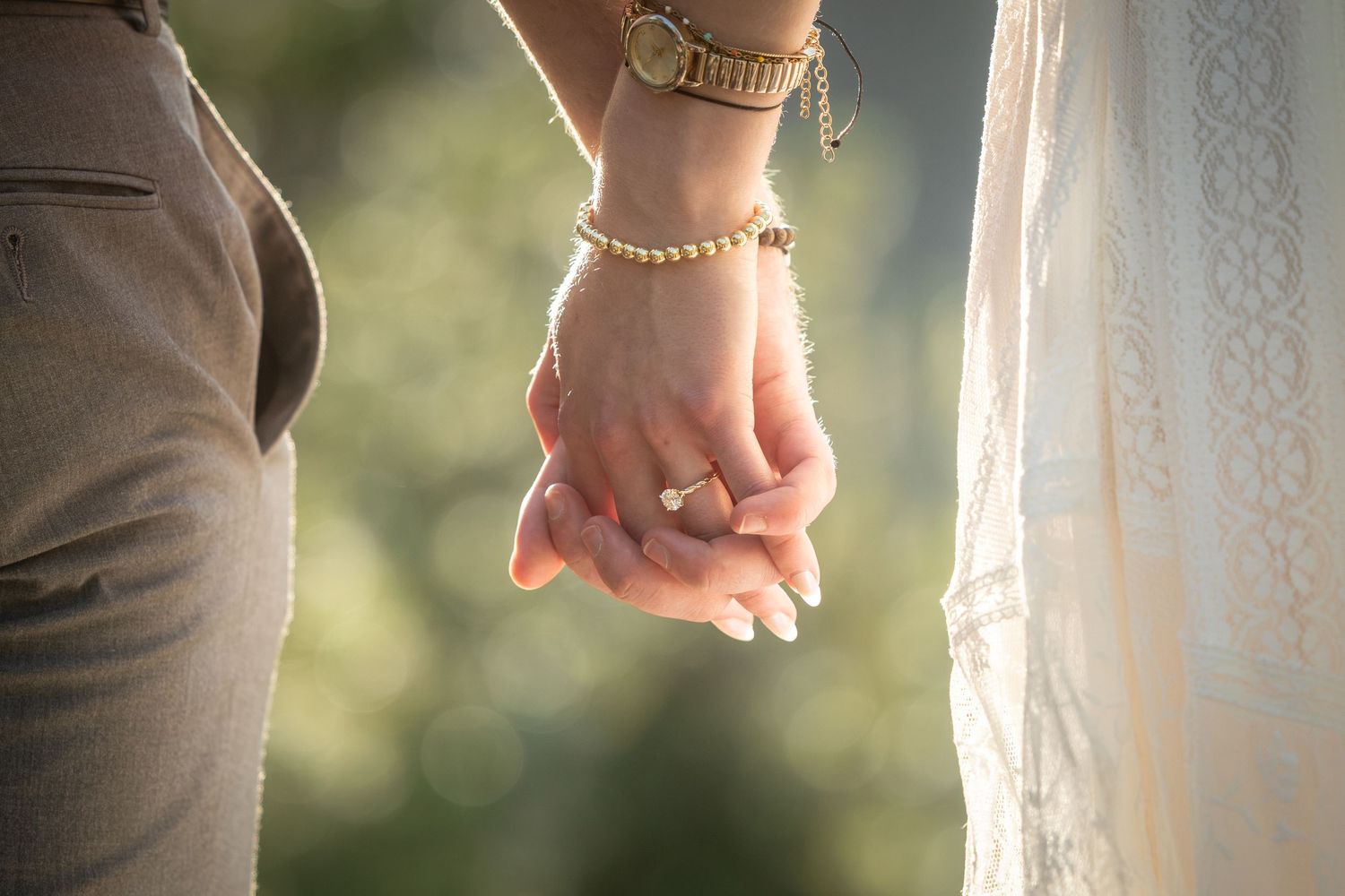 Close-up of a bride and groom holding hands, showcasing a gold bracelet and wedding ring at an outdoor ceremony.