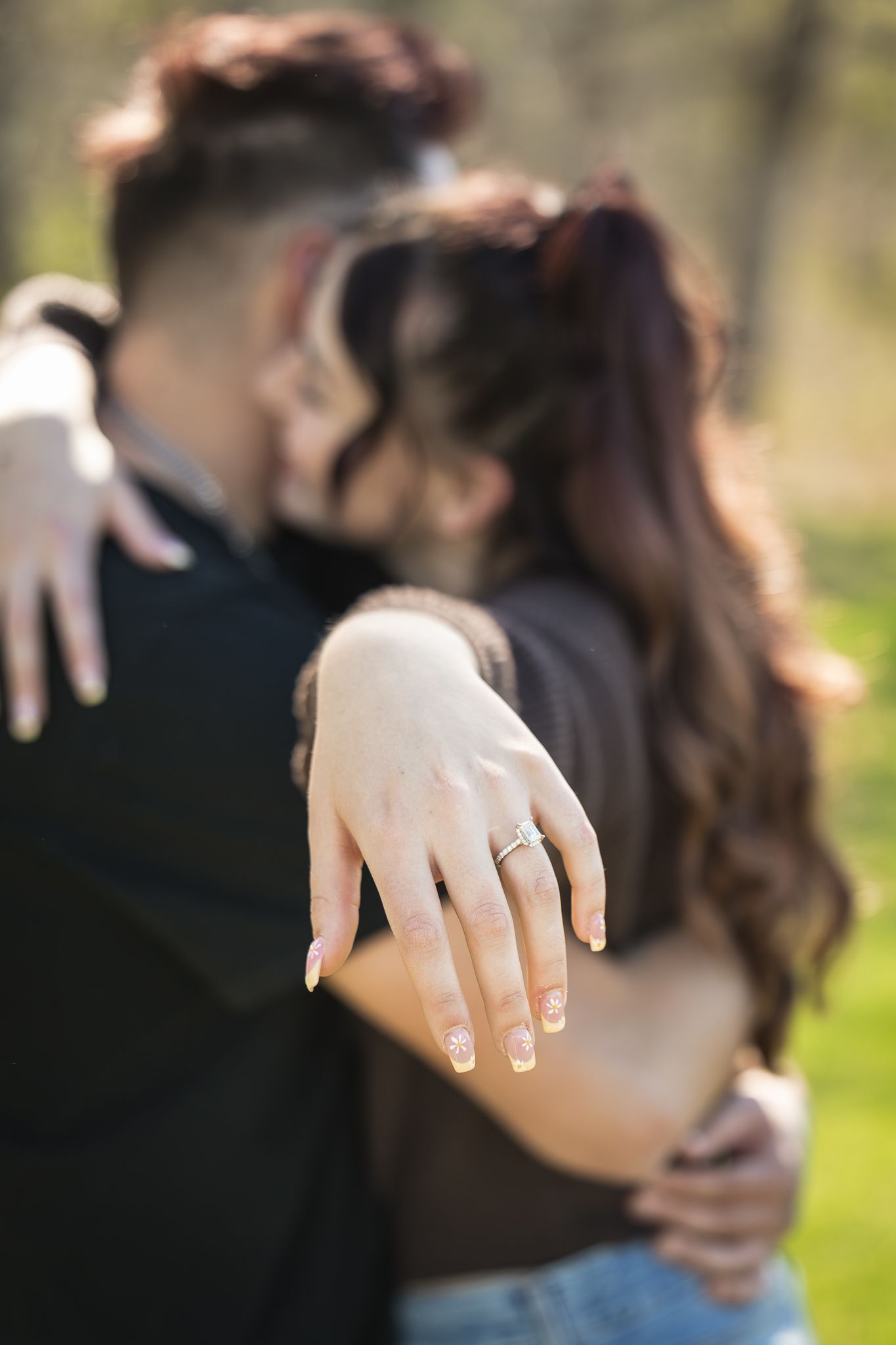 Couple embracing outdoors while woman displays engagement ring on her finger in sunny park setting.