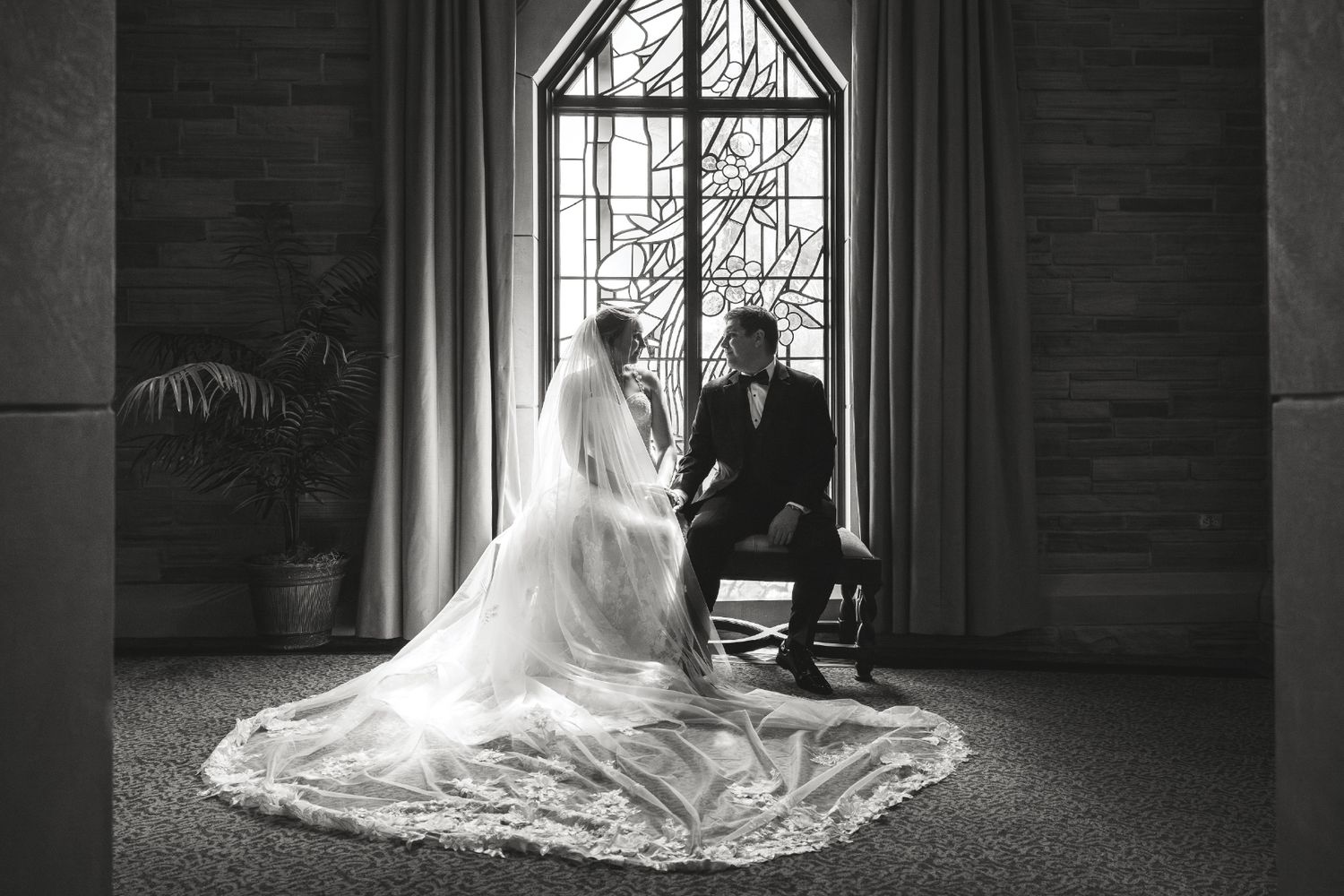 Black and white Colorado wedding photo of bride and groom sitting by stained glass window, long lace veil trailing on floor.