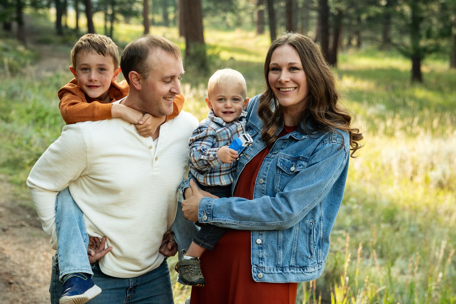 Happy family of four posing outdoors in a forest setting, parents holding two young boys, smiling warmly.
