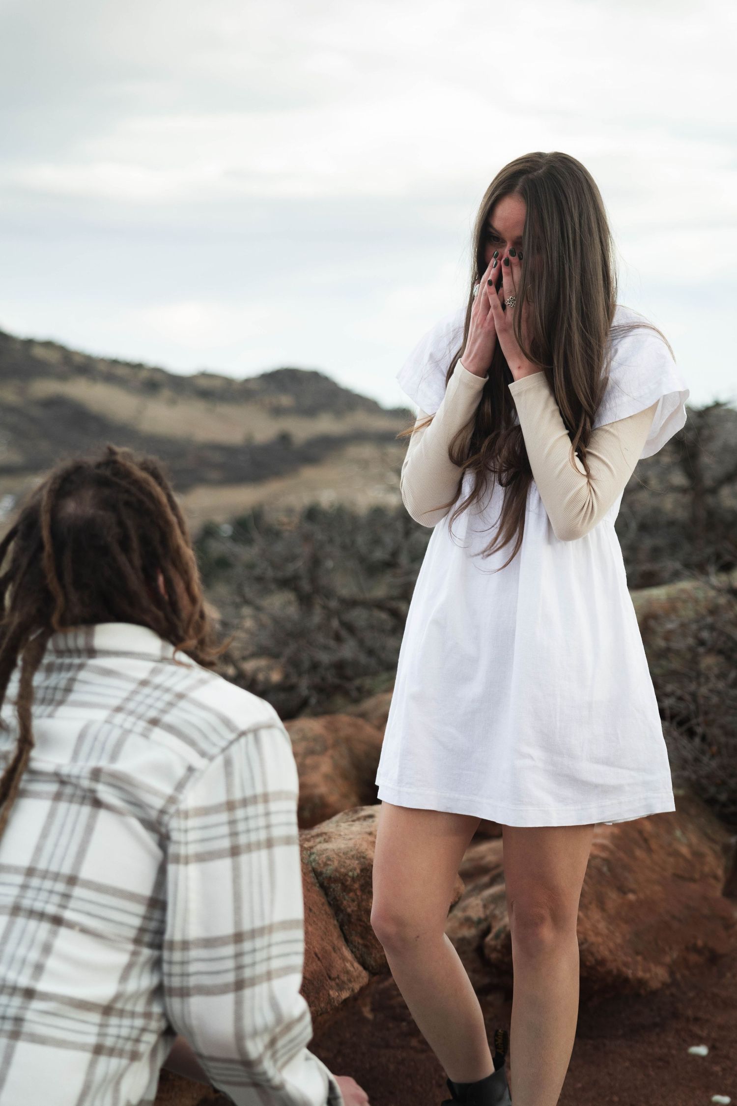Person in white dress reacts emotionally while another kneels on rocky terrain during a surprise moment.