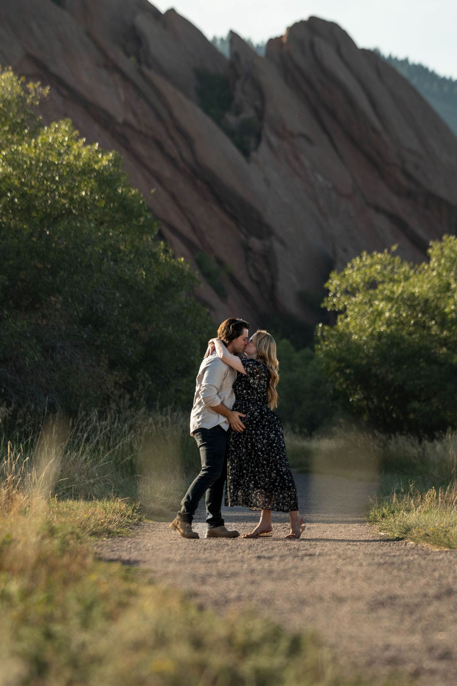 A couple embraces on a trail with dramatic red rock formations in the background.