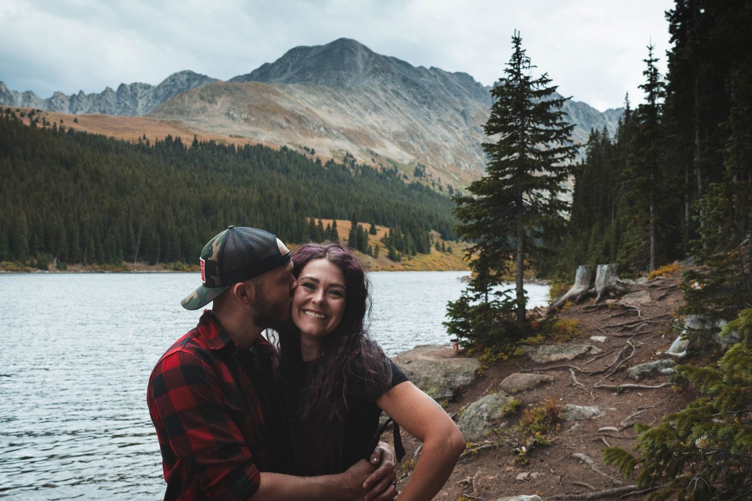 Couple embracing on rocky shoreline with mountain lake and evergreen forest in background.