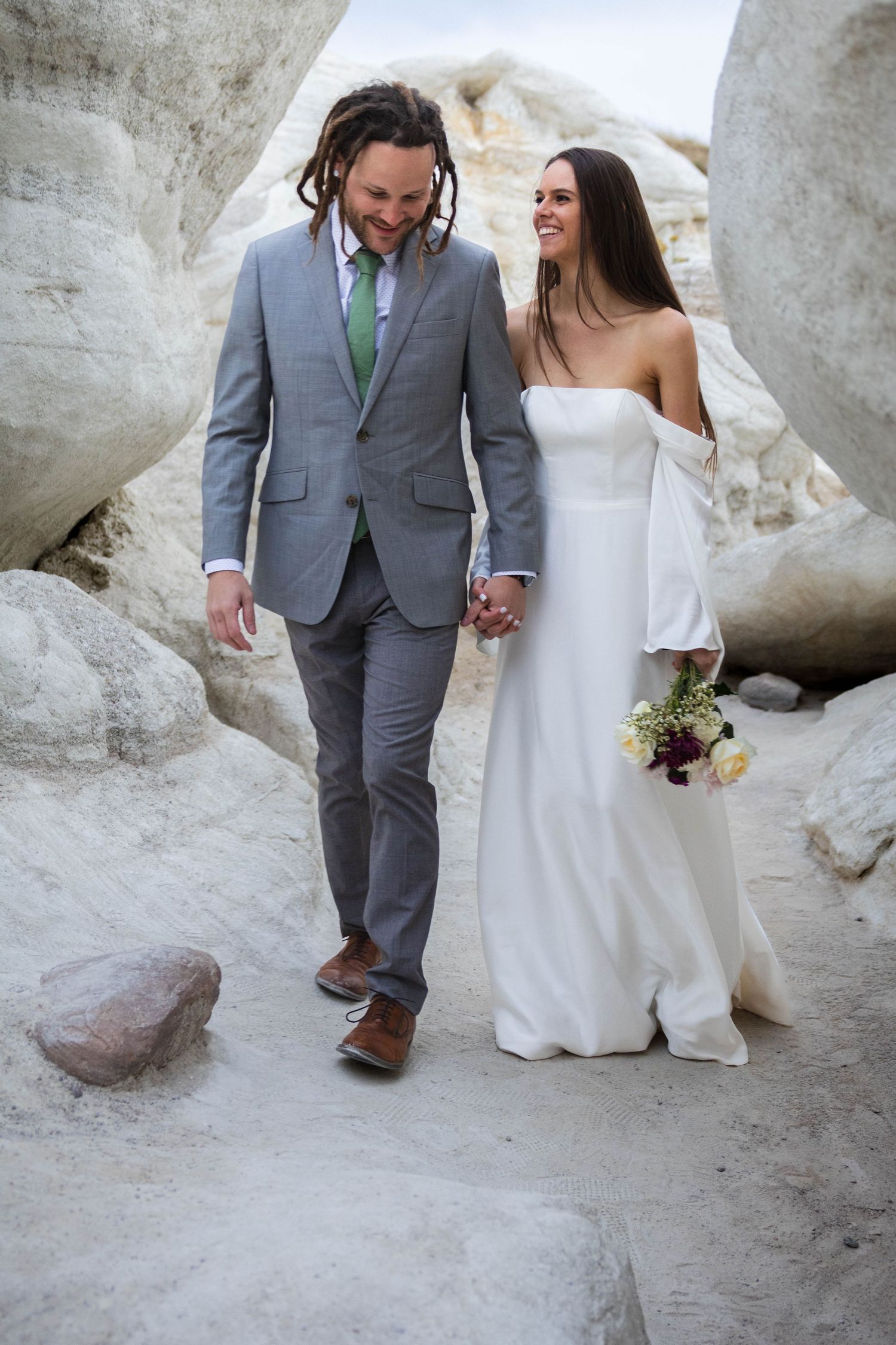 A couple explores rocky desert terrain on their wedding day, dressed in formal attire.