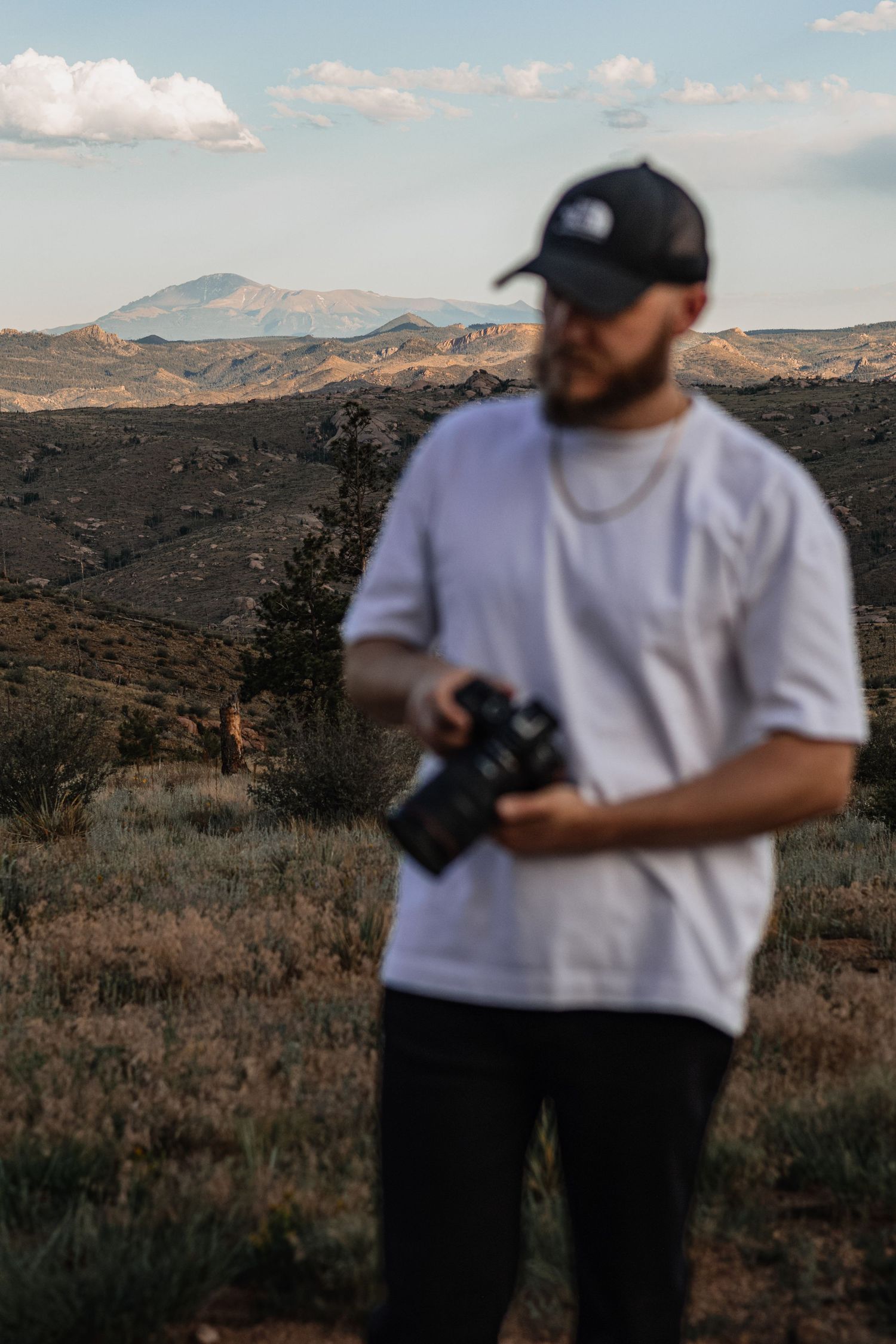 A photographer in white t-shirt and baseball cap captures desert landscape views against a mountainous backdrop at sunset.