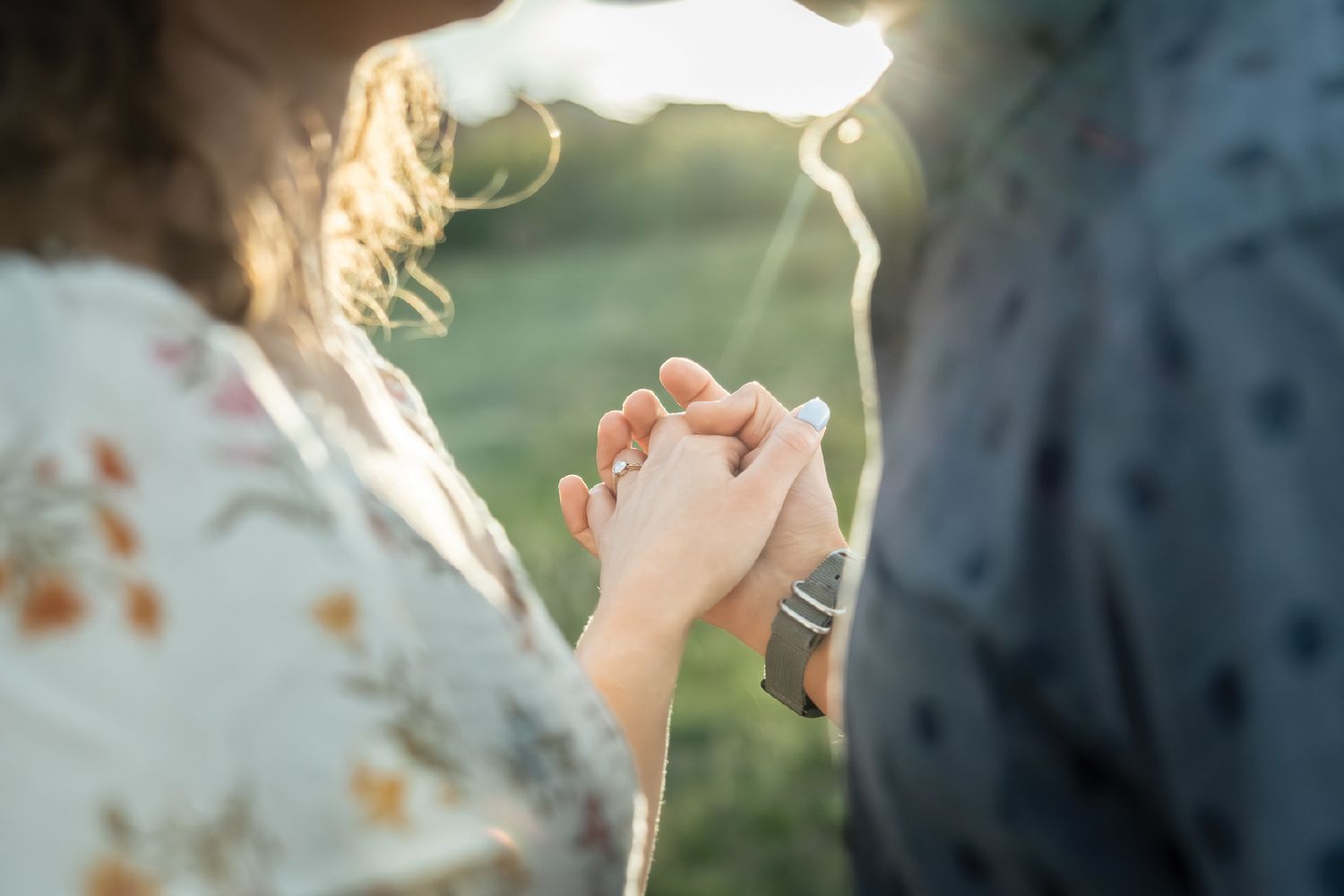 Two people holding hands outdoors in golden sunlight, one wearing a floral dress and the other a watch.