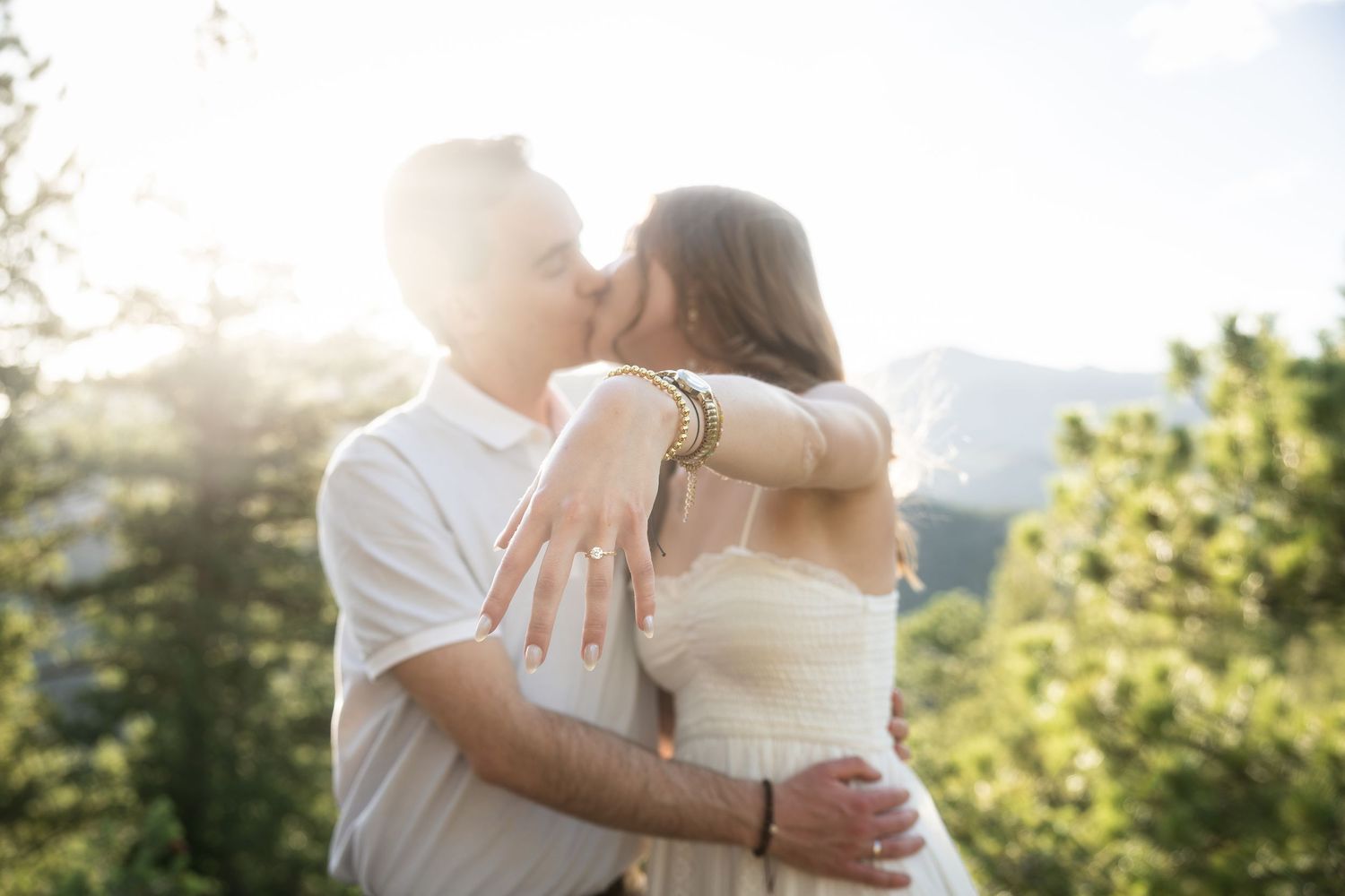 Couple sharing a romantic kiss outdoors with sunlight and mountain views, woman showing engagement ring.