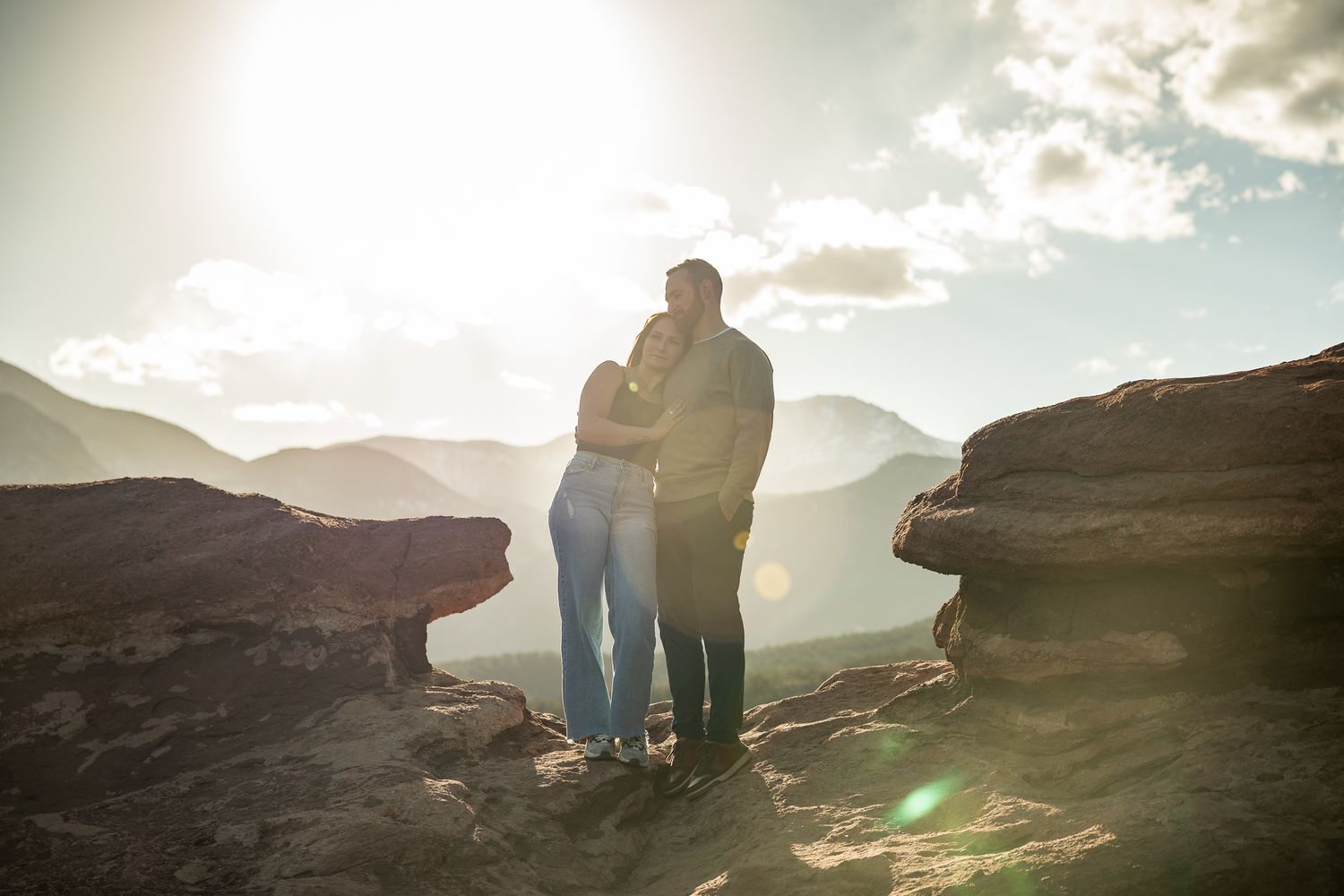 Couple embracing on rocky mountain summit at sunset with scenic valley and glowing sunlight behind them.