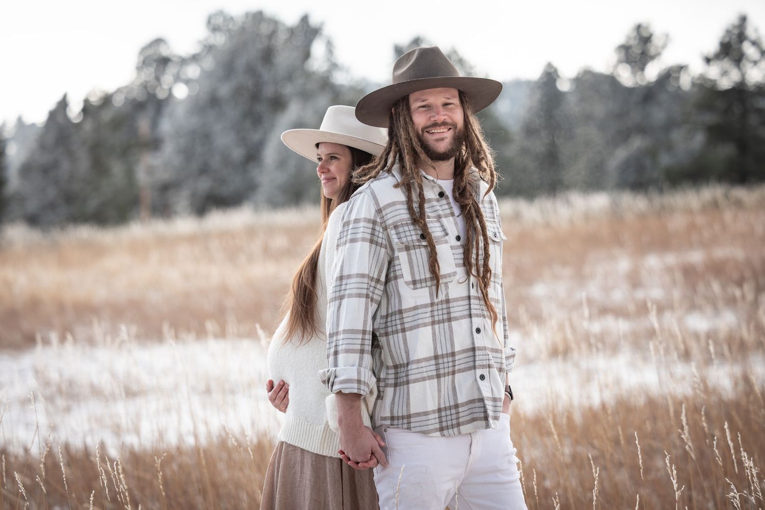 Couple in wide-brim hats holding hands in a snowy field, man with dreadlocks smiling in plaid shirt.