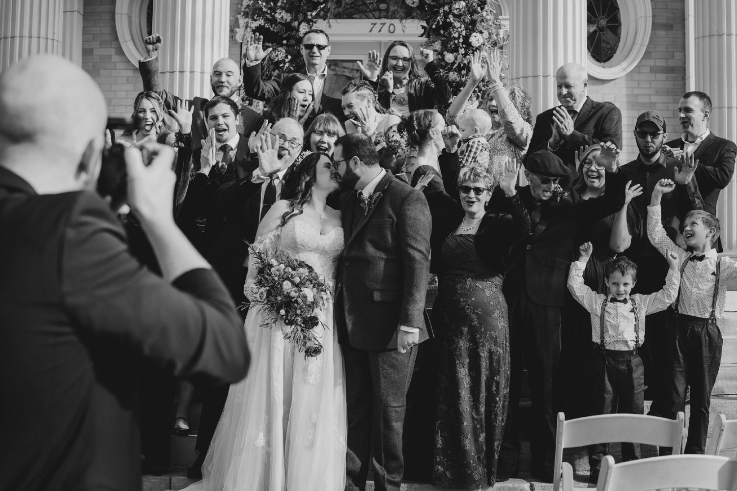 Black and white wedding photo of couple kissing surrounded by cheering guests on church steps.