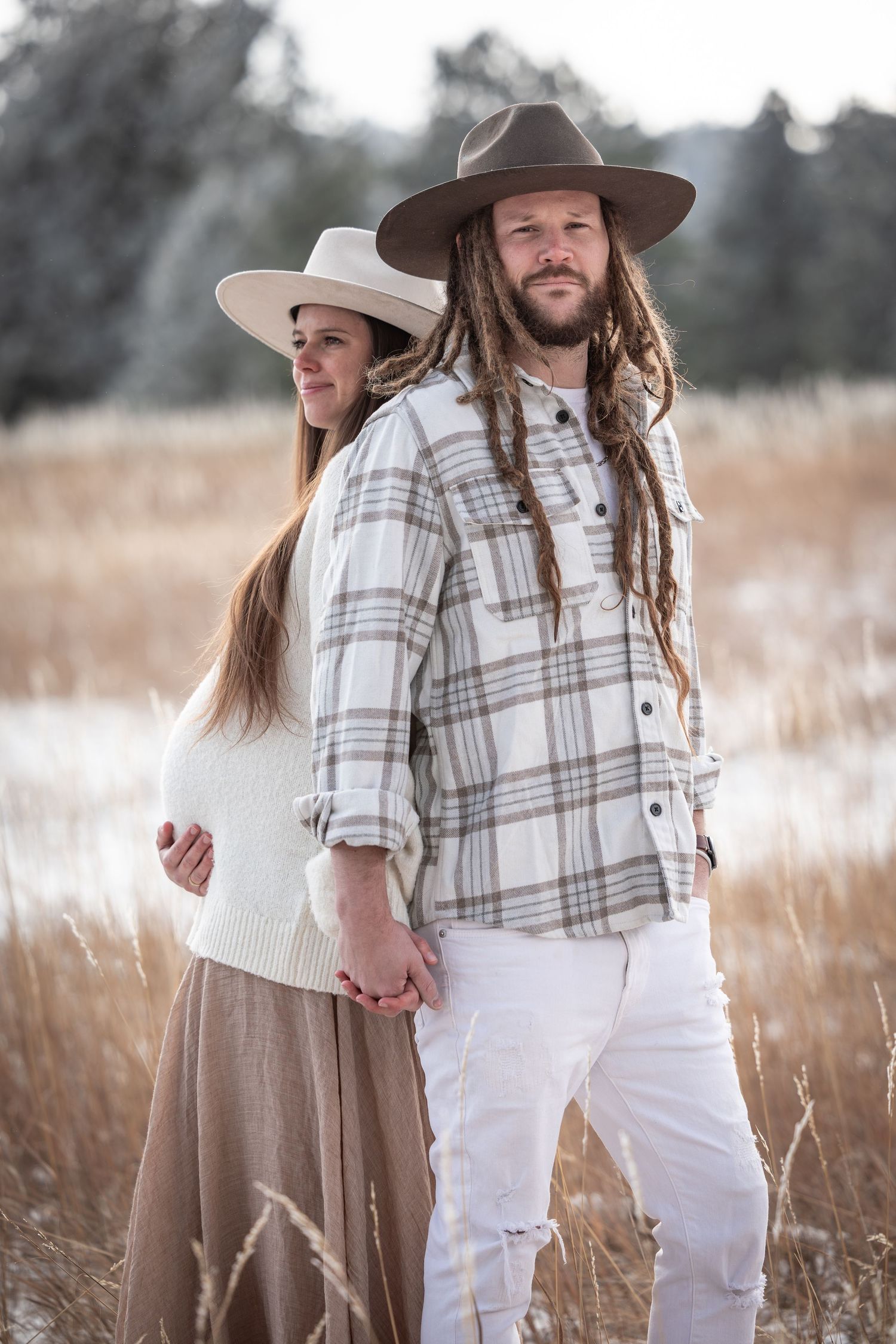Couple in coordinated neutral outfits with plaid and brown hats pose together in a rustic autumn field.